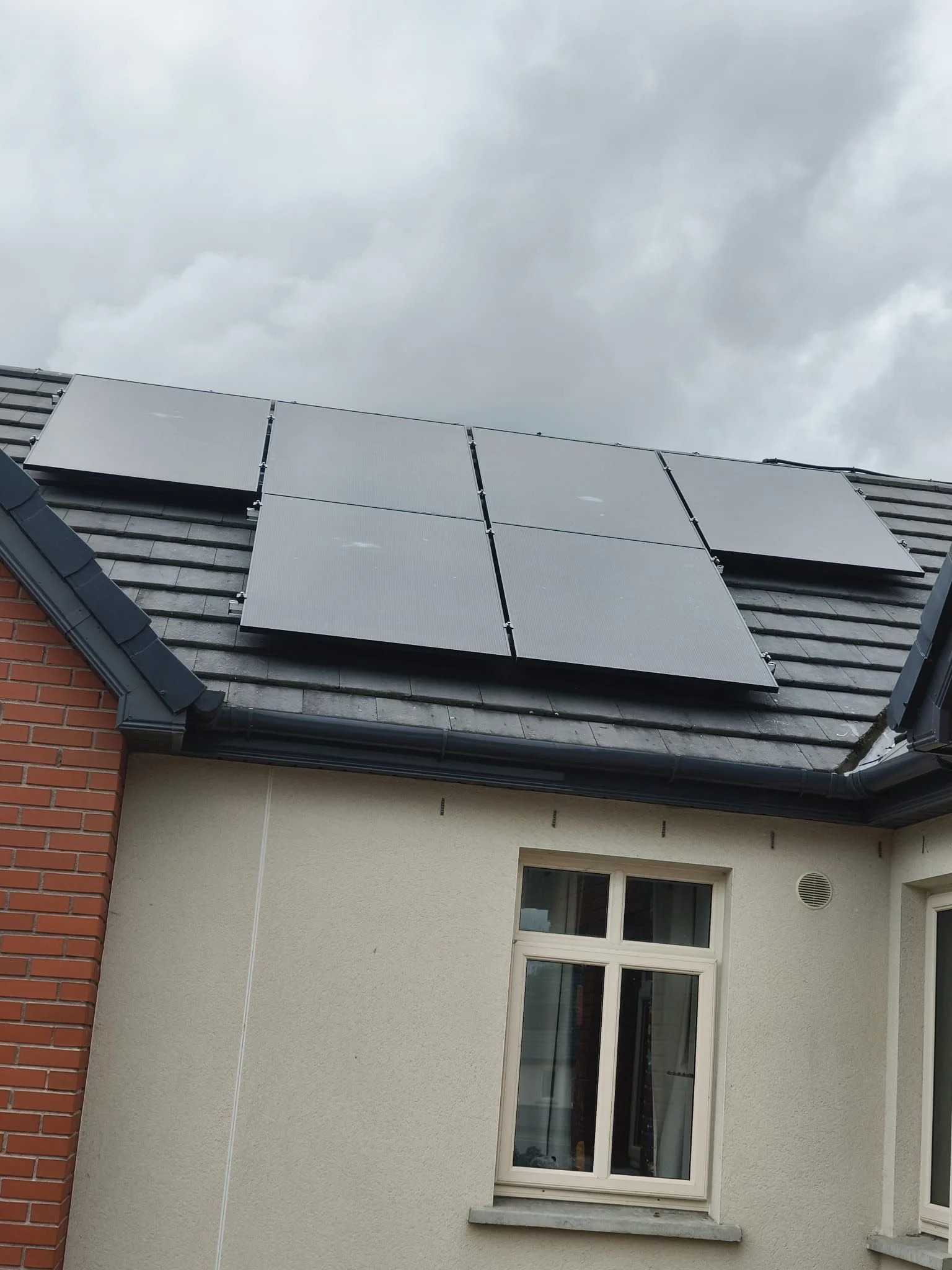 Roof with solar panels on a cloudy day, part of a house with a window and brick wall.