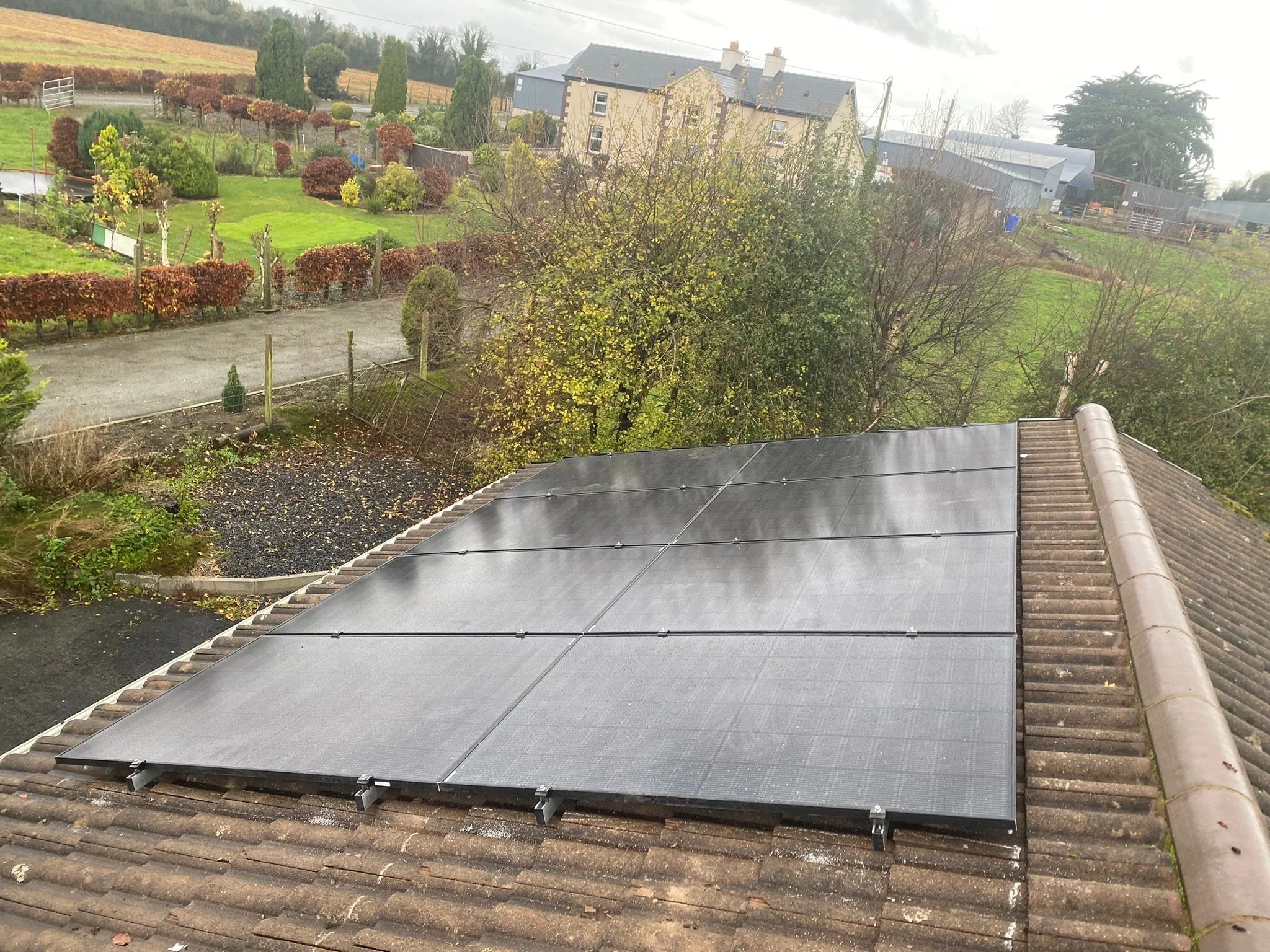 A rooftop with solar panels installed on brown tiles, overlooking a garden with trees and a lawn, and a rural landscape in the background.