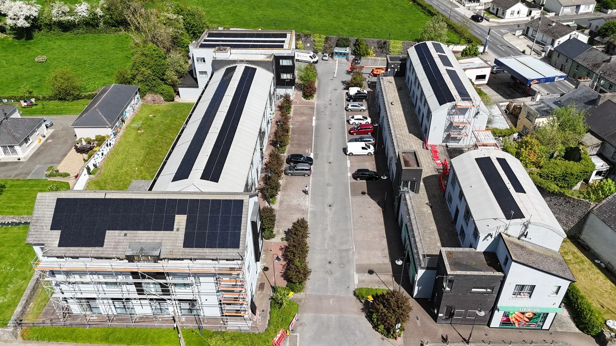 An aerial view of a street with commercial and residential buildings, with solar panels on the roofs, parked cars, trees, and a grassy area with a playground.