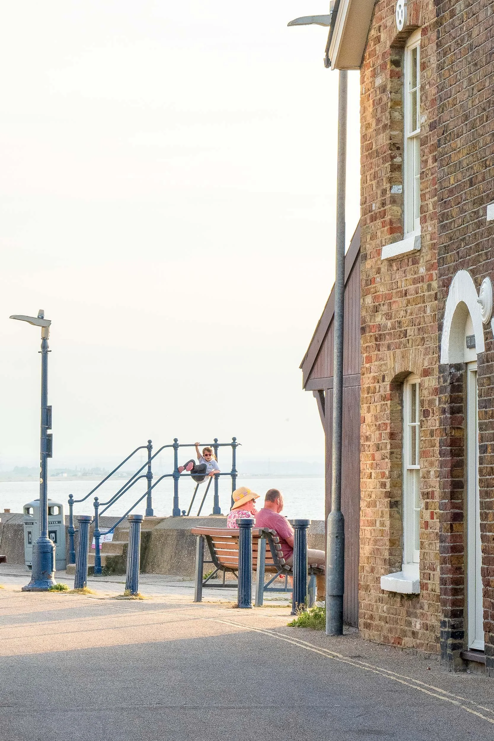 People sitting on a bench near a brick building along a waterfront, with a child playing on a railing in the background and a waterfront view beyond. Whitstable, Kent, England.