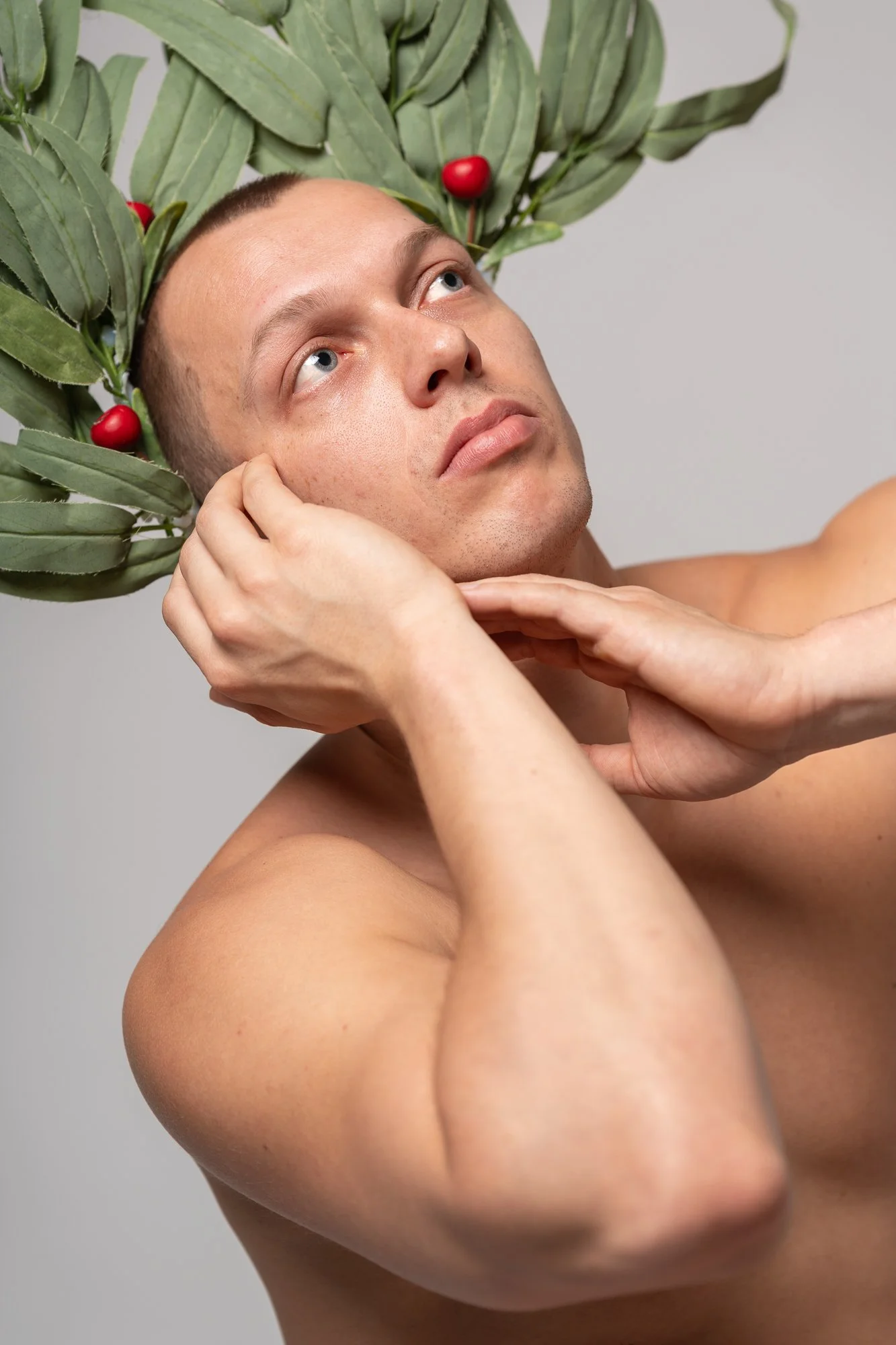 A shirtless man with a decorative leafy crown with red berries, looking upward with one hand on his face and the other supporting his chin.