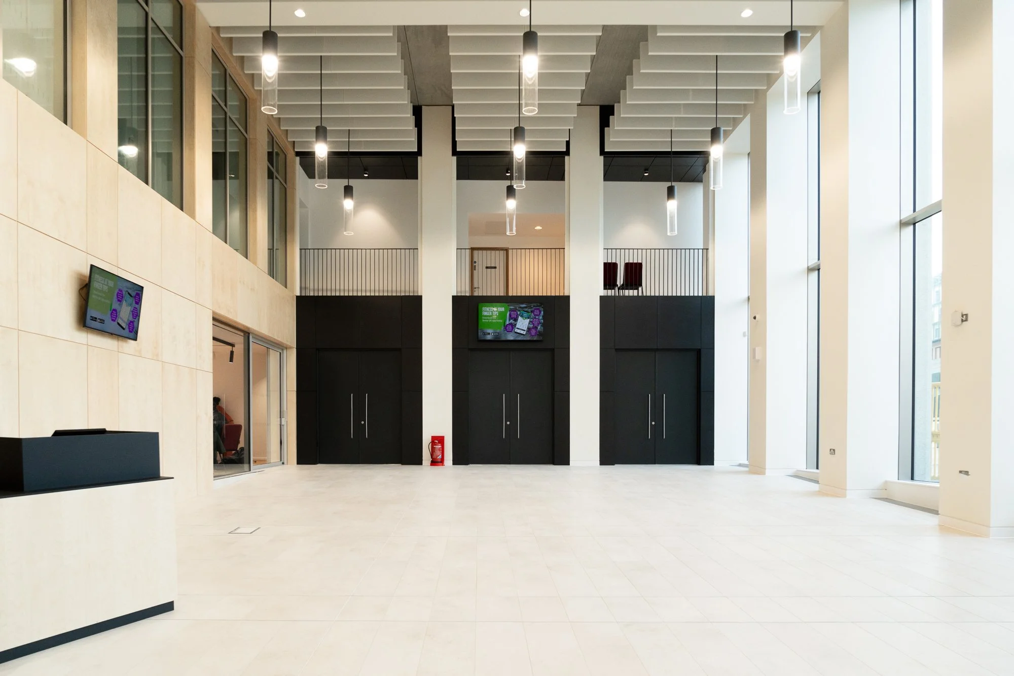 A spacious, modern lobby with large windows, high ceilings, hanging lights, black and white walls, and a digital screen. There are two levels with a staircase and railings, with an empty floor space.