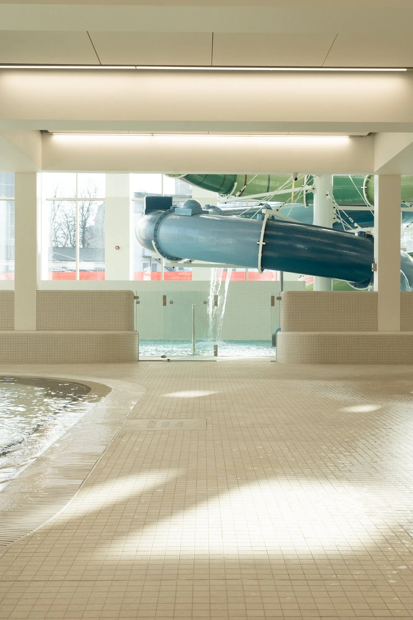 Indoor water park with a large twisting water slide, water pouring out at the end, and a pool below, with large windows in the background letting in natural light.