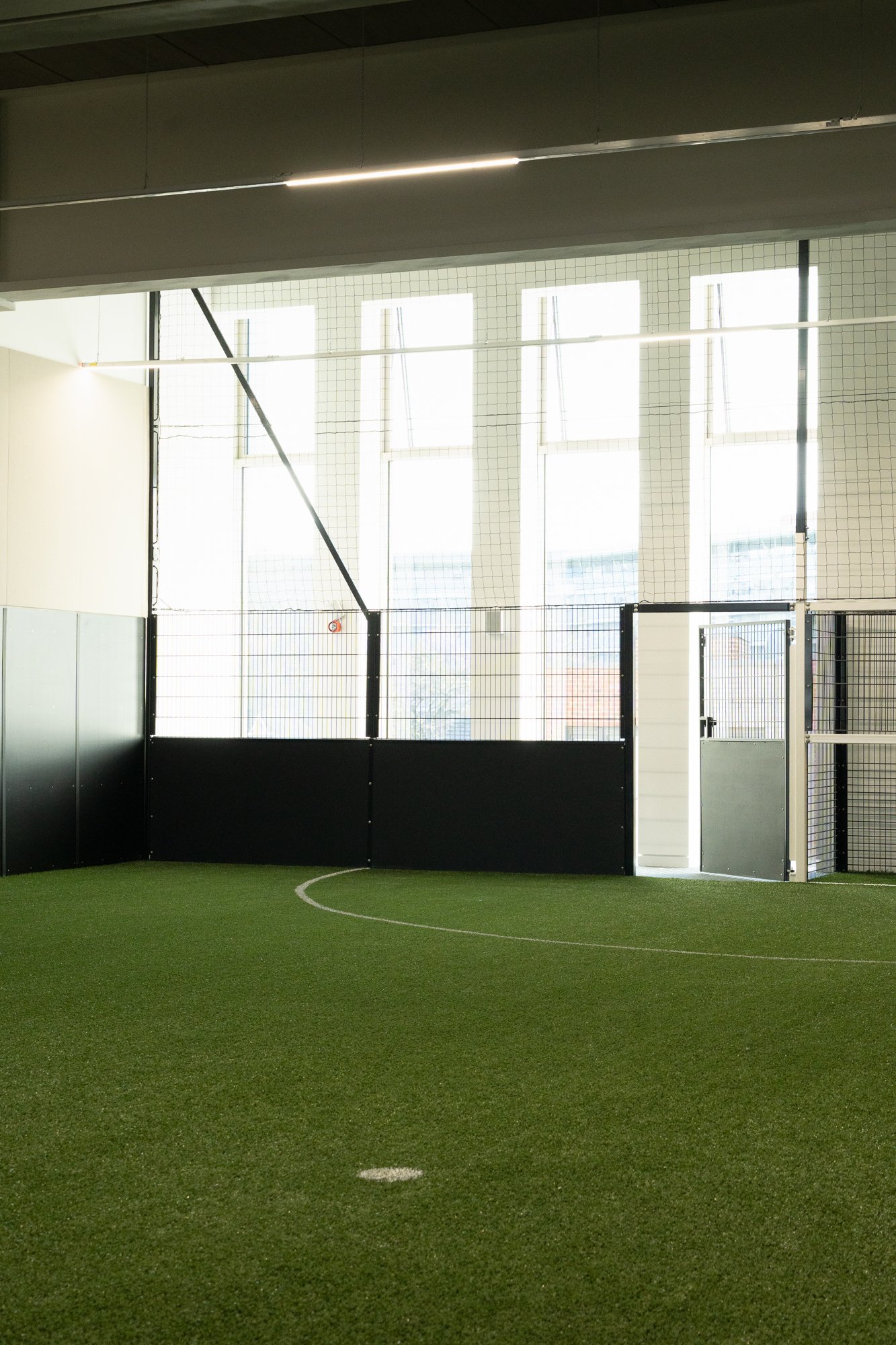 Indoor soccer field with artificial turf, surrounded by high windows and black fencing, with a door on the right side.