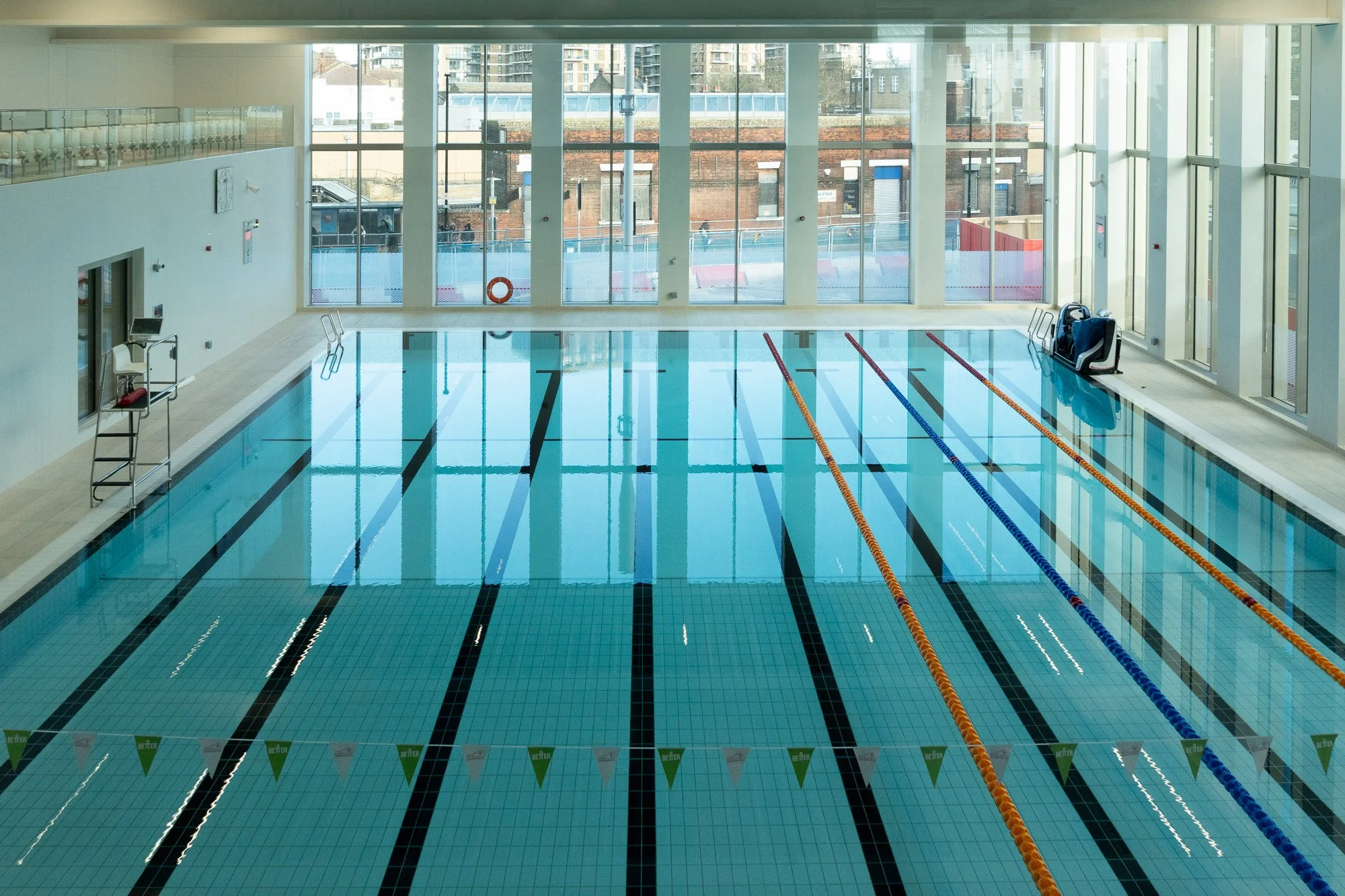 Empty indoor swimming pool with lane markers, large windows, and outdoor cityscape view.