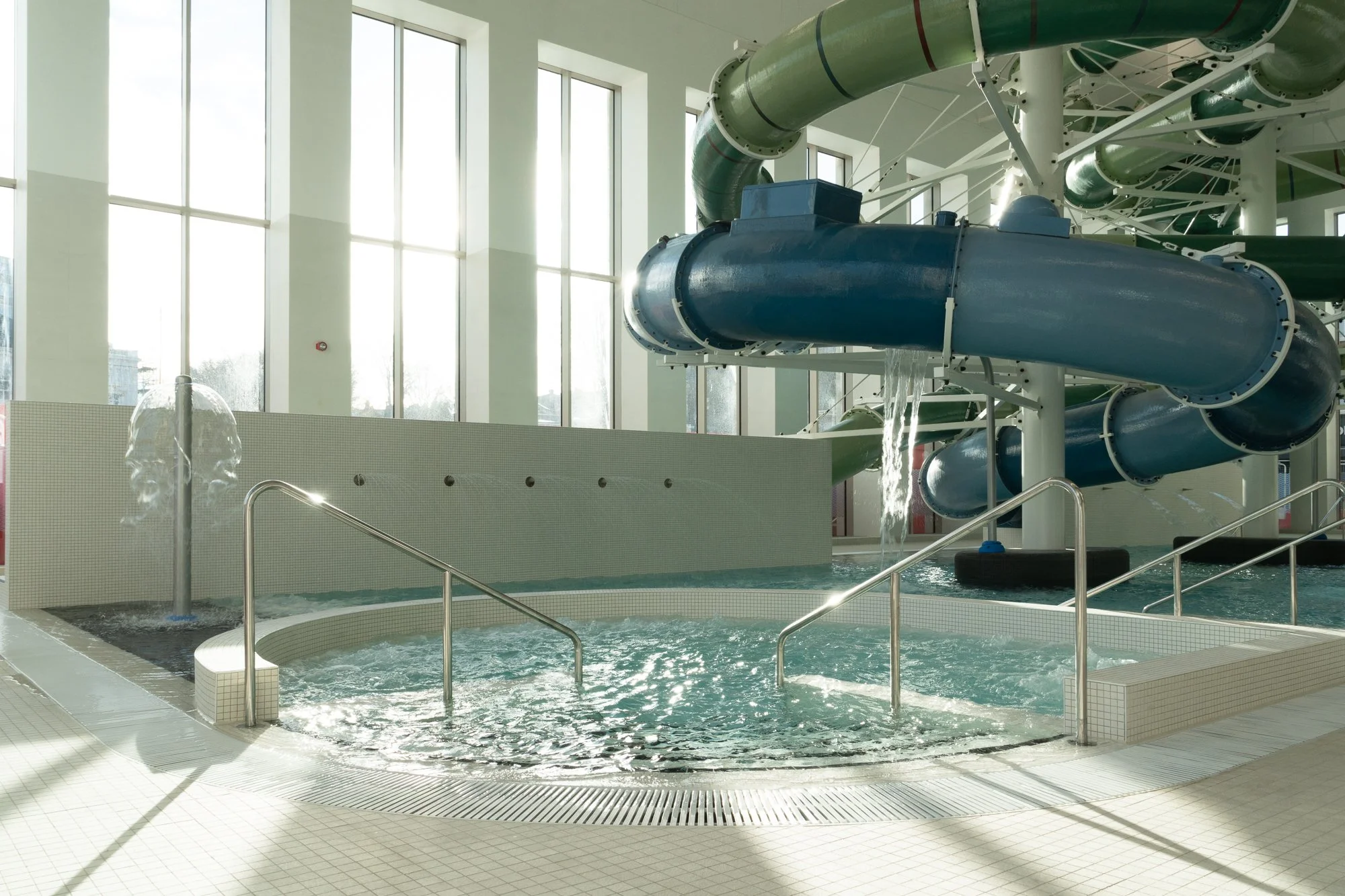 Indoor water park featuring a large blue water slide, hot tub with metal railings, and a water fountain in front of tall windows letting in natural light.