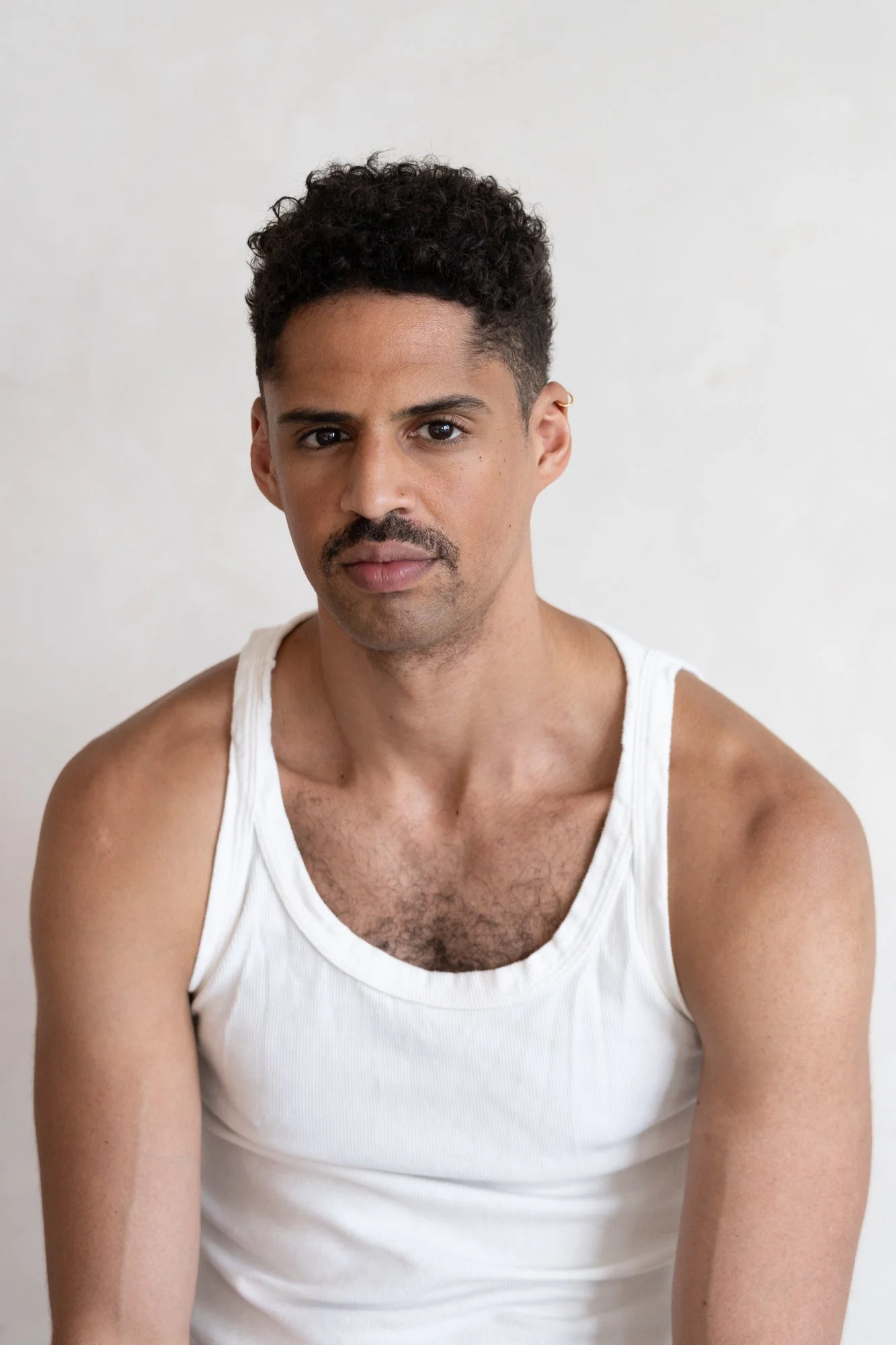 A man with curly black hair, mustache, and medium skin tone wearing a white tank top, sitting against a plain white background.