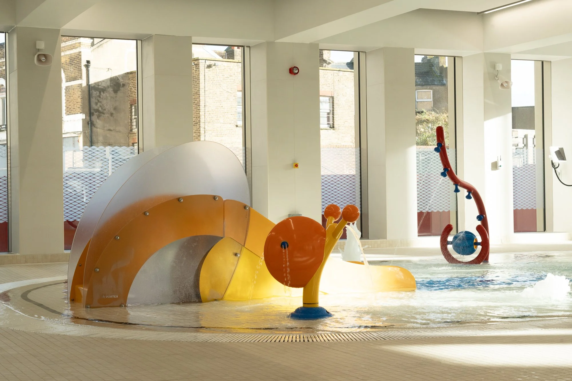 Indoor children’s water play area with colorful water features, including a snail-shaped fountain and a curved water slide, set near large windows.
