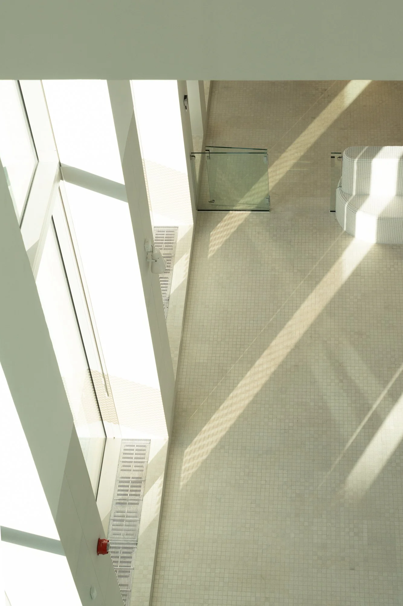 Bright indoor space with large glass windows, tiled white floor, glass door, and seating area with curved white bench.
