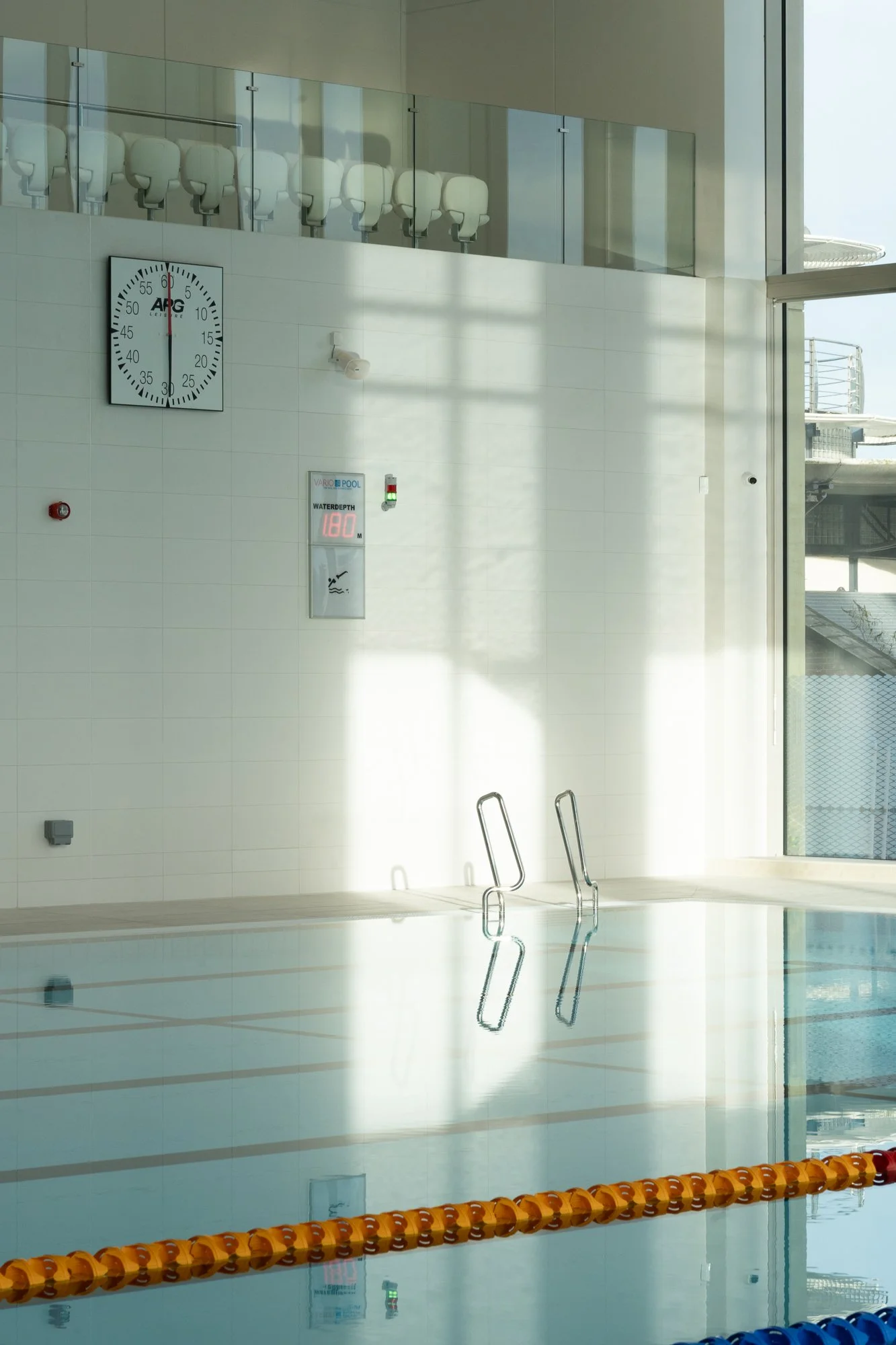 Indoor swimming pool with a ladder, a clock on the wall, and sunlight streaming through large windows.