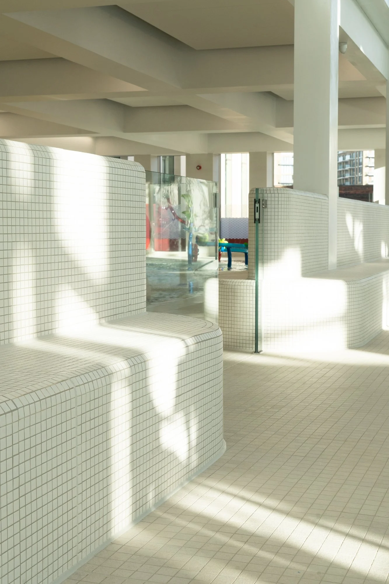 Light-filled indoor pool area with white tiled walls and floor, glass partitions, and a view of the pool with slides and toys in the background.
