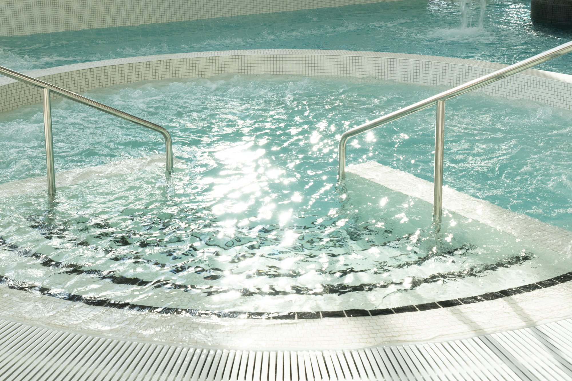 A hot tub with stainless steel handrails, filled with bubbling water, in an indoor spa or pool area.