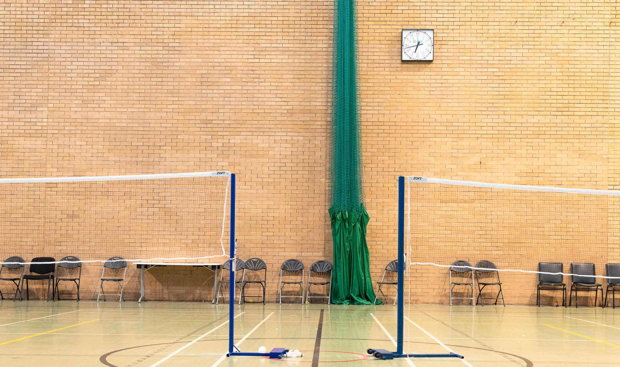 Indoor gymnasium with badminton courts, chairs along the wall, a clock on the brick wall showing 2:20, and a green curtain hanging from the ceiling. Waterfront Leisure Centre, Woolwich, London, England.