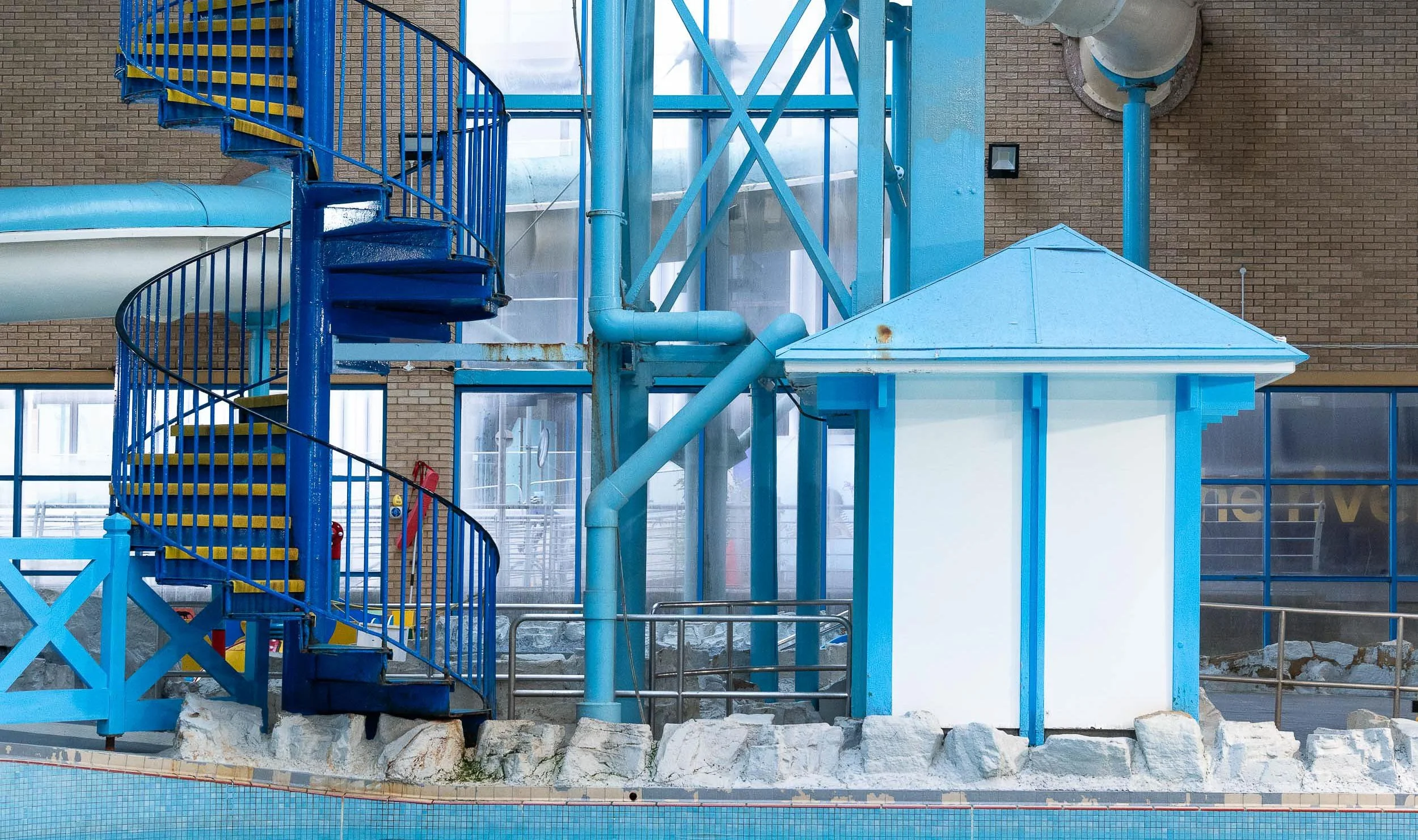 Indoor water park feature with blue spiral staircase, water slide, water pipes, and mineral rocks at the base. Waterfront Leisure Centre, Woolwich, London, England.