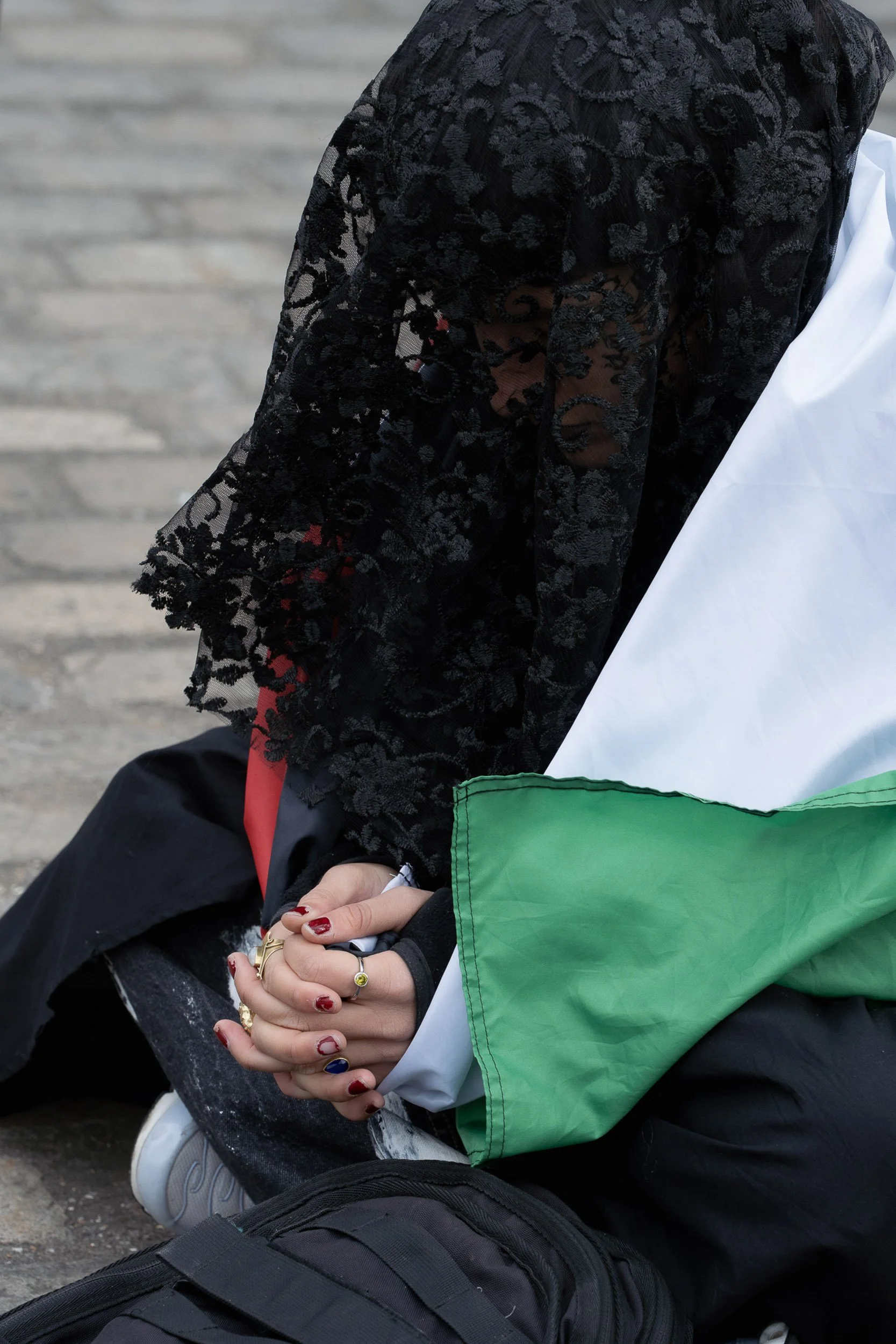 Person sitting on the ground with hands clasped, wearing a black lace top, white and green jacket, black pants with white paint stains, and white shoes, with a black backpack nearby.