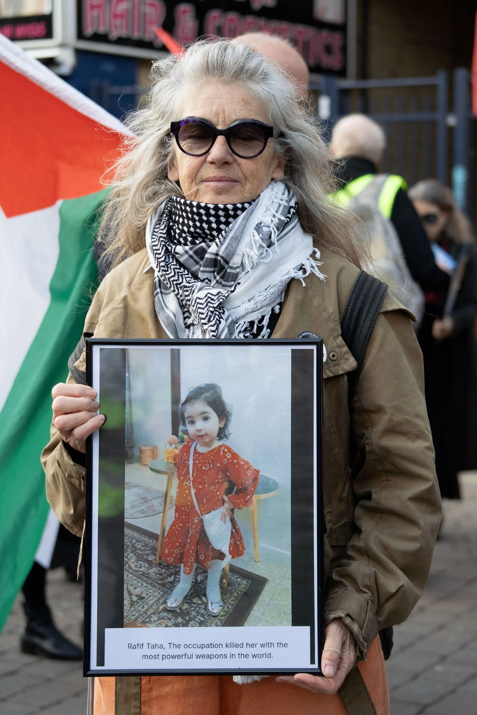 An older woman with gray hair, sunglasses, and a patterned scarf holding a framed picture of a young girl in an orange dress. The frame has a caption reading: 'Rafif Taha, The occupation killed her with the most powerful weapons in the world.' In the