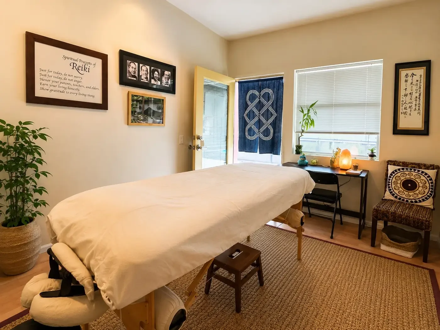 A massage or therapy room with a massage table covered in a white sheet, a small stool, a chair with a decorative pillow, a plant by the window, and framed pictures and artwork on the wall.