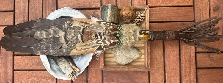 Decorative Native American ceremonial staff with feathers, a carved wooden bear head, and a stone, against a wooden background.