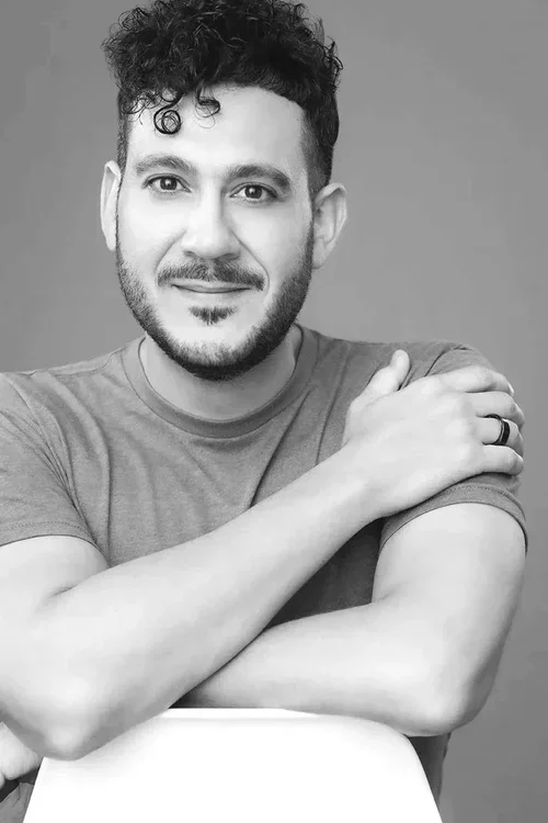 A black and white portrait of a young man with curly hair and a beard, smiling slightly, wearing a t-shirt, with arms crossed in front of a plain background.