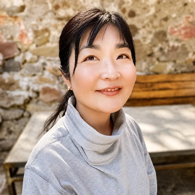 A smiling Asian woman with black hair tied back, wearing a light gray turtleneck, sitting outdoors against a stone wall and wooden bench.