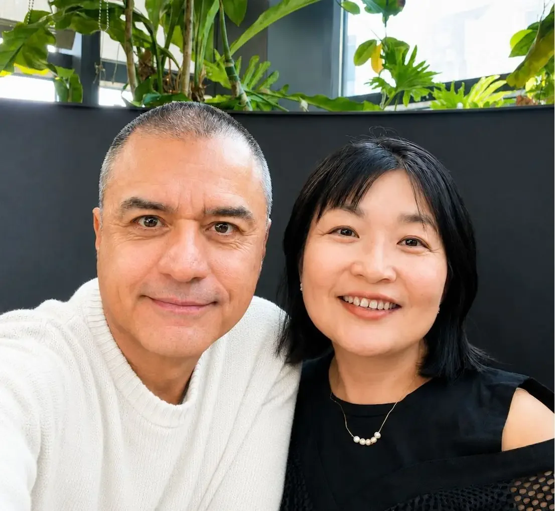 A man and a woman taking a selfie together indoors, with greenery in the background.