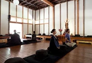 People meditating in a serene room with wooden walls, large windows, and a statue of a religious figure at the front.