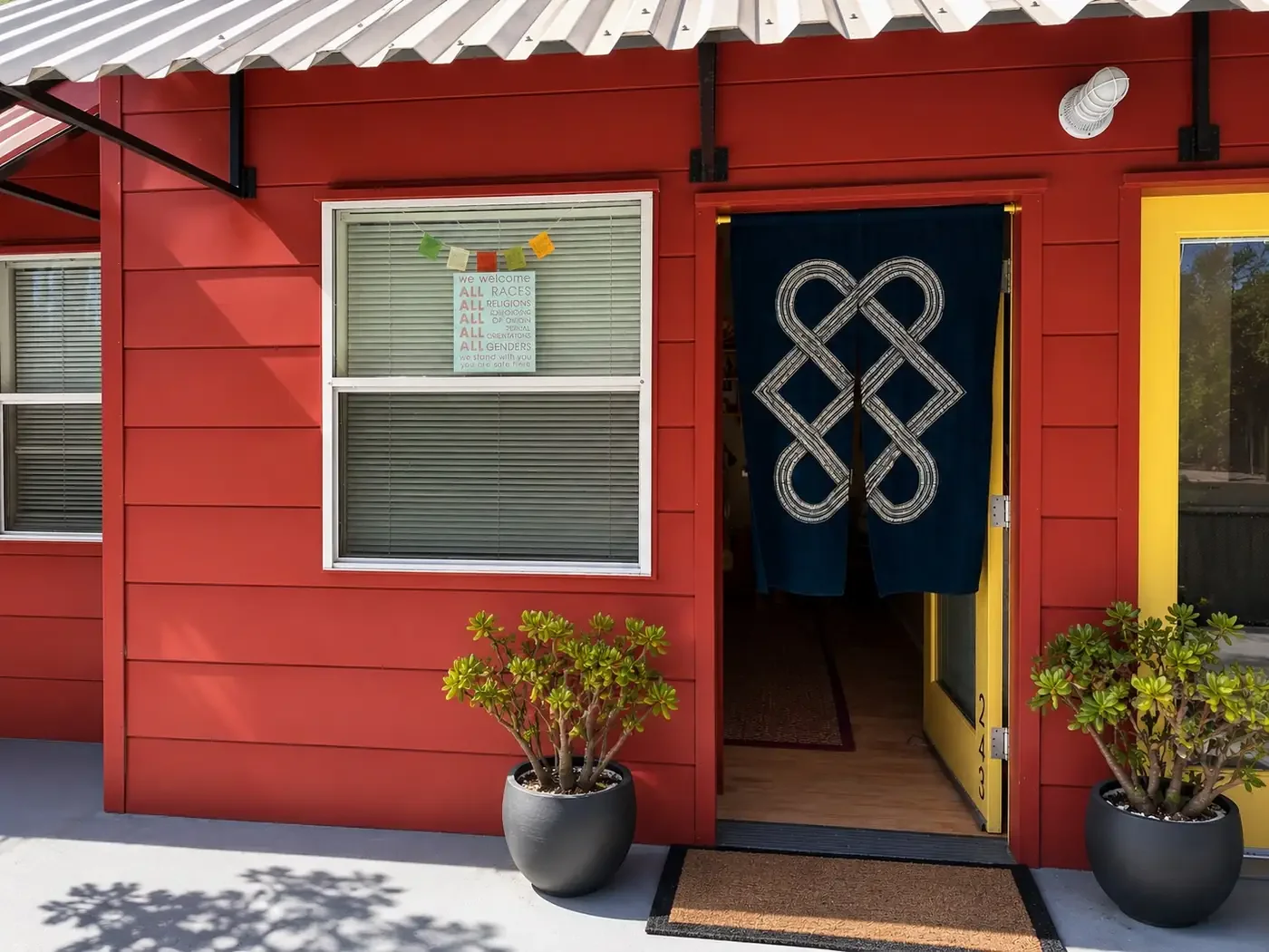 Red building with a blue cloth curtain with a white geometric design hanging over the doorway, potted plants on each side of the entrance, a sign in the window welcoming all races, religions, genders, and orientations, and a yellow door panel next to the entrance.