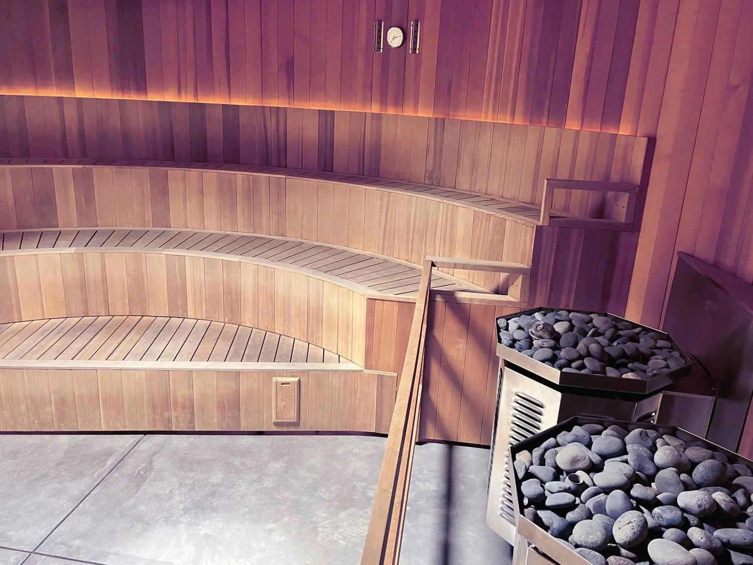 Inside a sauna with curved wooden benches and a heater filled with stones.