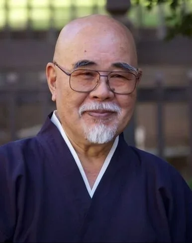 An Reiki sensei in Kyoto, Japan with glasses and a white beard, wearing traditional Japanese clothing, standing outdoors with a blurred background.