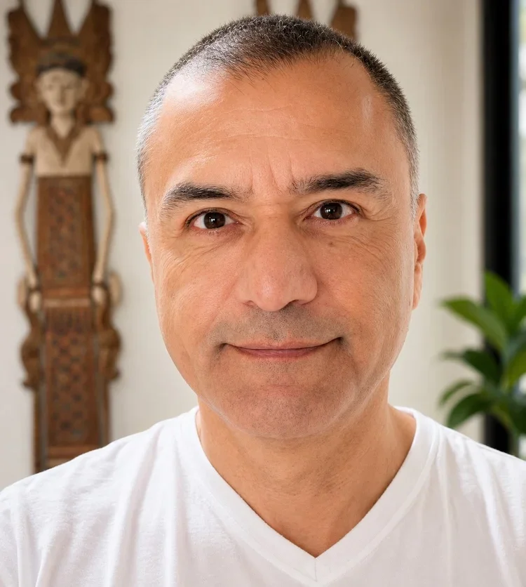Close-up of a middle-aged man with short hair, smiling, wearing a white t-shirt, indoors with a plant and a decorative wooden wall hanging in the background.