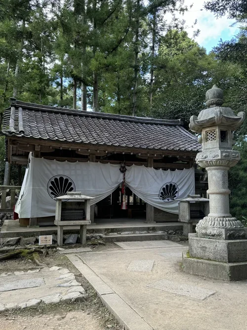 Small traditional Japanese shrine with a wooden structure, white curtain with a black fan symbol, stone lantern, and surrounded by trees.