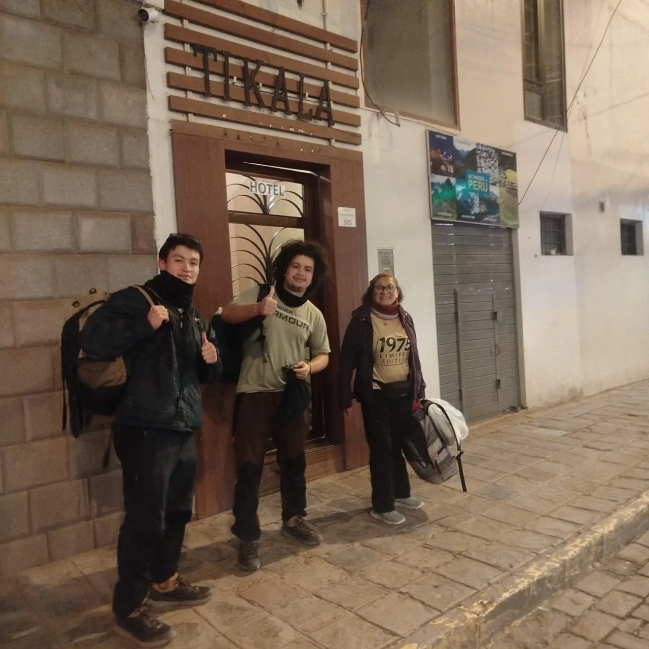 Tres personas con mochilas en la calle frente a un hotel en una noche, sonriendo y haciendo señal de capitán con los pulgares.