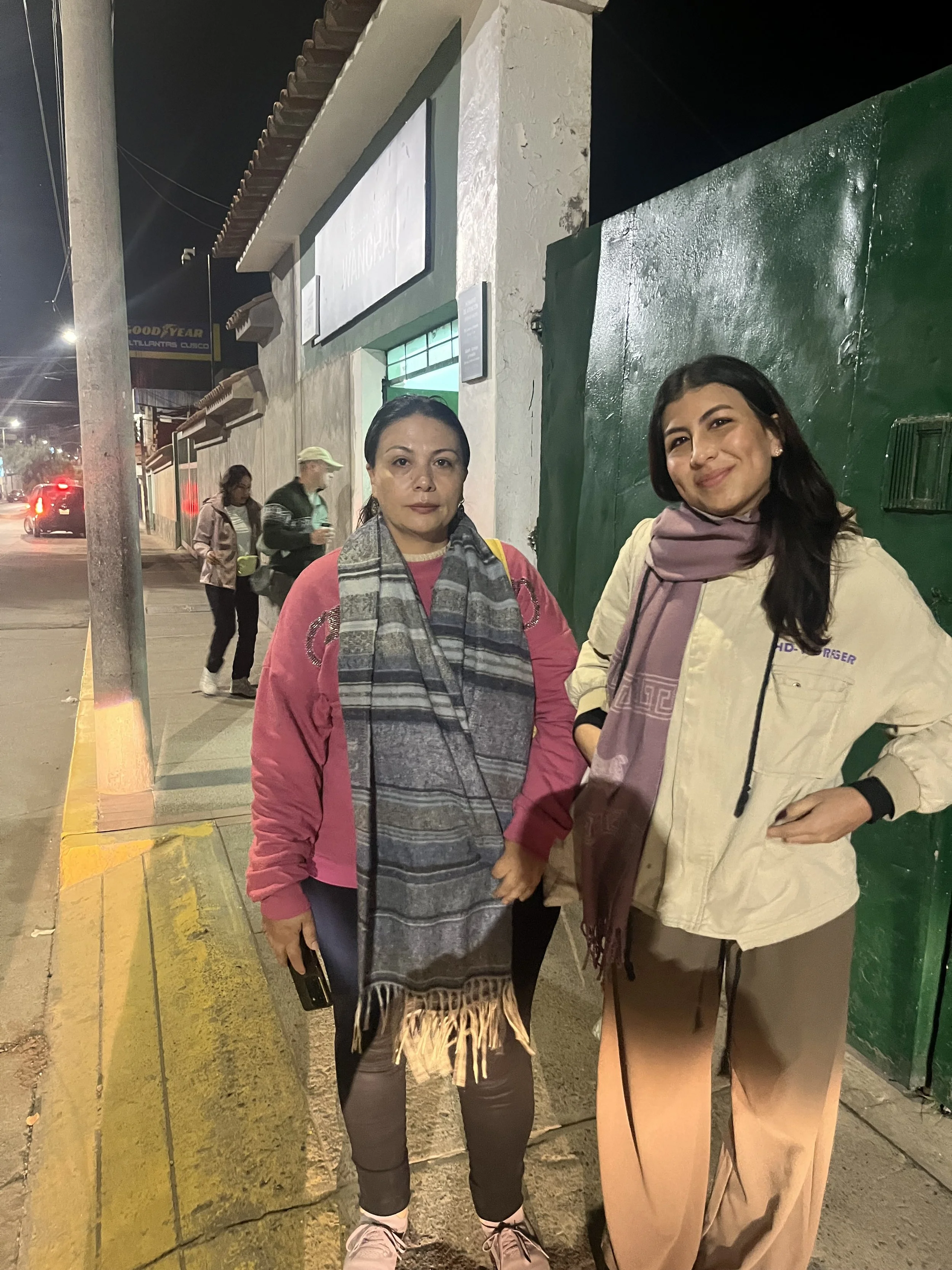 Dos mujeres de pie en la acera durante la noche, en un entorno urbano con un edificio blanco y verde al fondo, y otras personas caminando en la calle.
