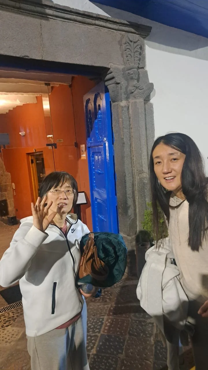 Dos mujeres de pie frente a una entrada de piedra con detalles tallados, en un interior con paredes de color naranja y piso de mosaico, una de ellas hace un gesto con la mano y la otra sostiene una chaqueta blanca.