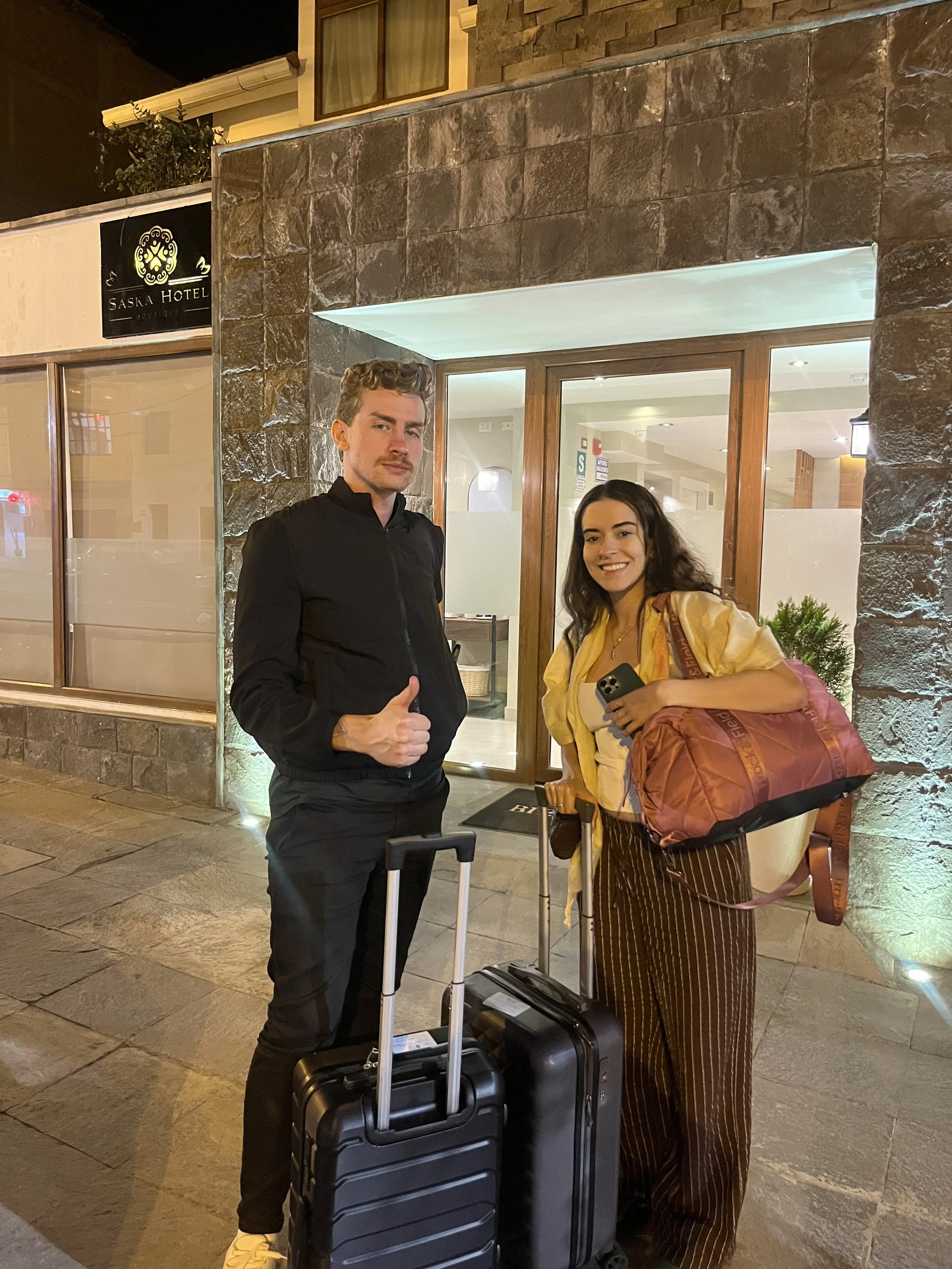 Dos personas con equipaje de mano en la entrada de un hotel, en la noche, sonriendo y con maletas.