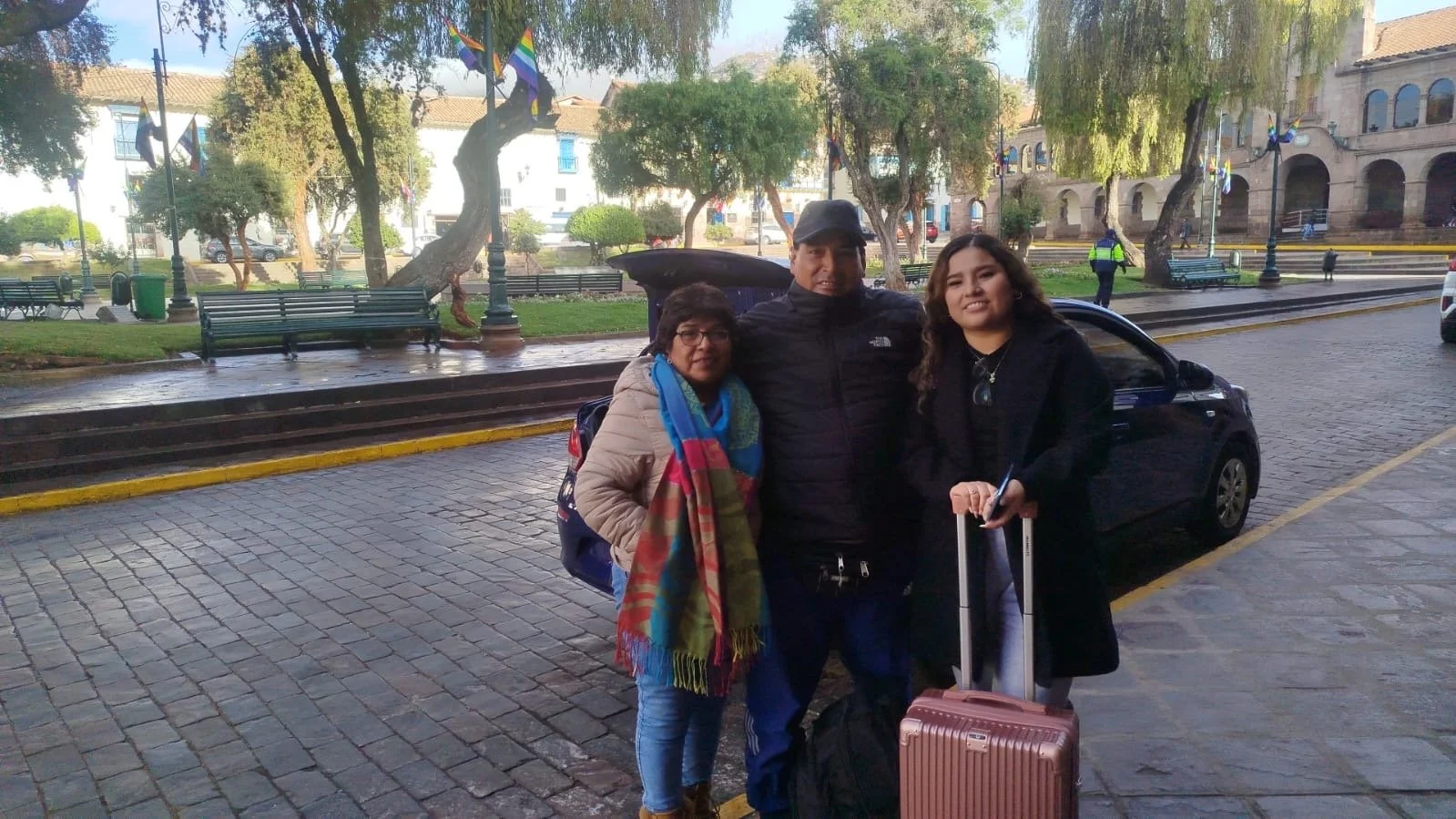 Tres personas en una calle empedrada con árboles y bancas, una sostiene equipaje de mano, otra lleva mochila y bufanda, y hay banderas en los árboles.