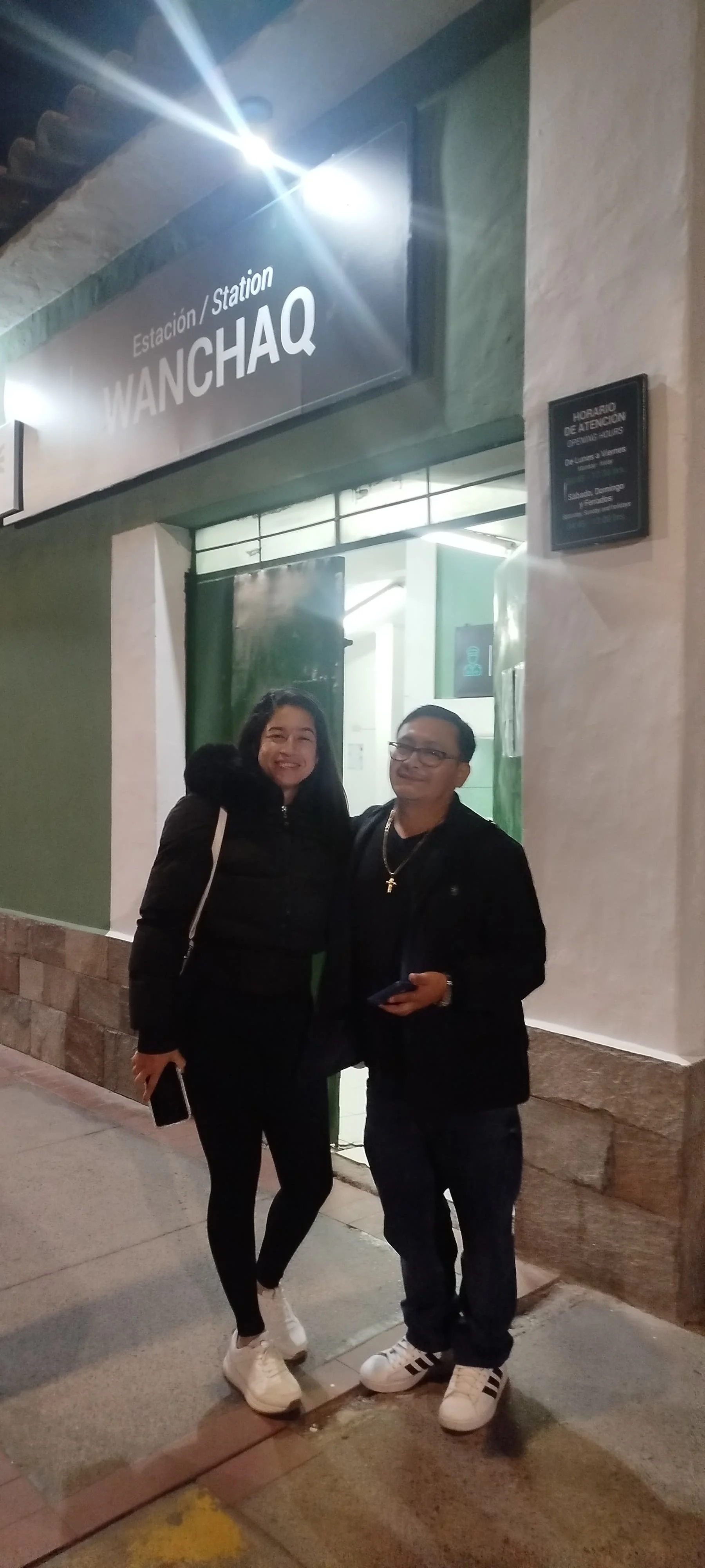 Dos personas, una mujer y un hombre, posando frente a la entrada de la estación de Wanchai, en la noche, con sonrisa, vestimenta casual y oscuras, la mujer con chaqueta negra y la mujer con un crucifijo, señalando la entrada.