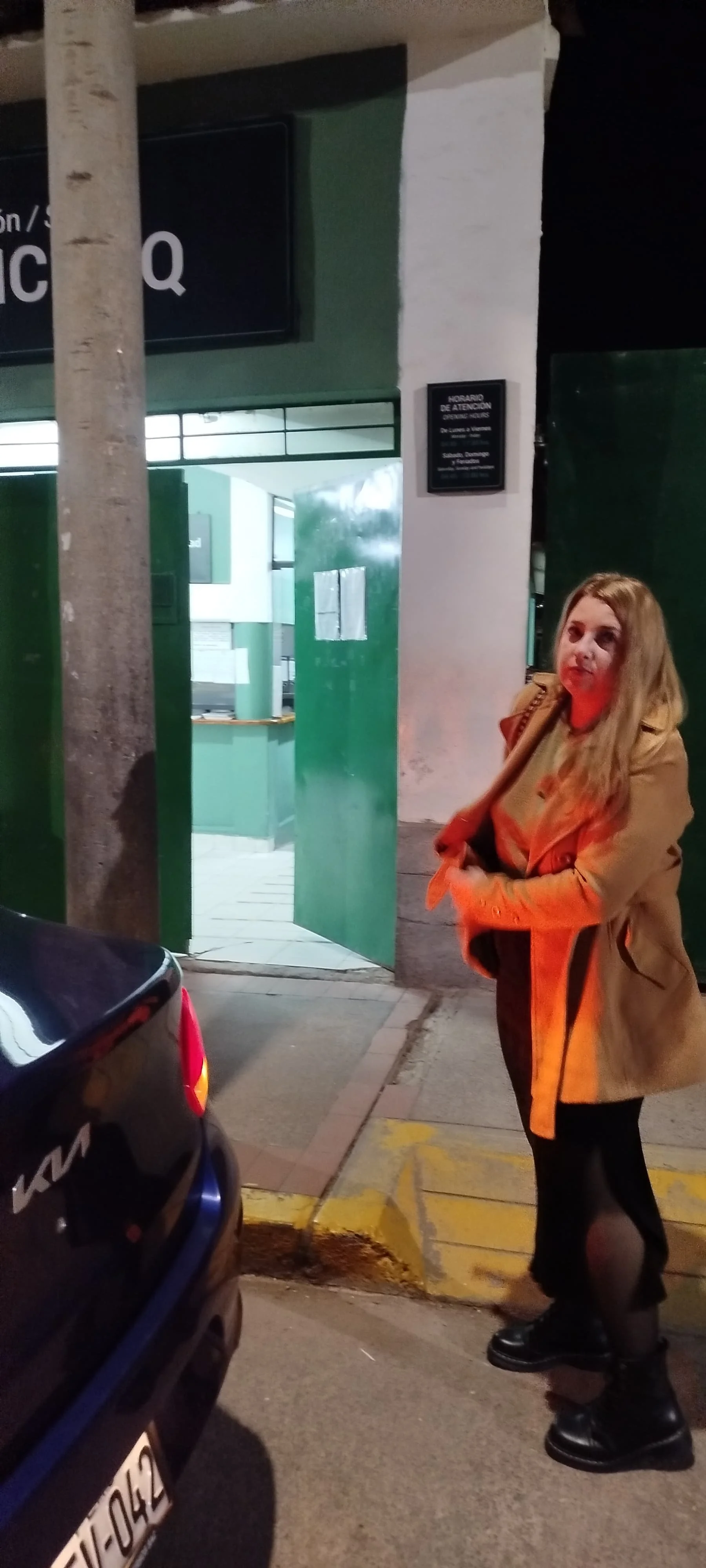 Mujer con chaqueta color beige y pantalones oscuros, de cabello rubio, de pie en la acera frente a la entrada de un establecimiento con puertas abiertas de color verde, en una calle, noche.