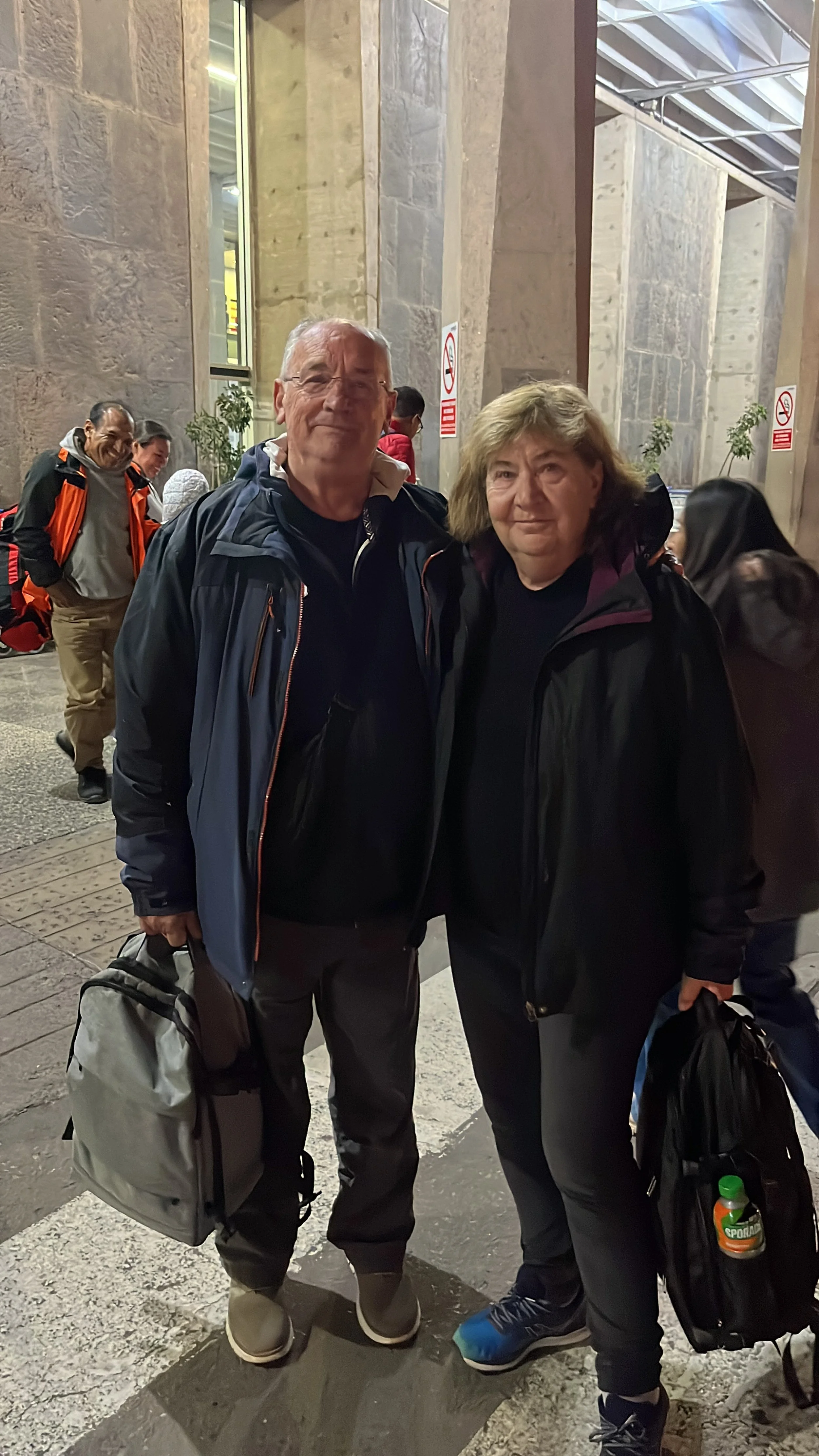 Dos personas con ropa de abrigo, sosteniendo bolsas de viaje, posando en una estación o terminal, con otras personas en el fondo.