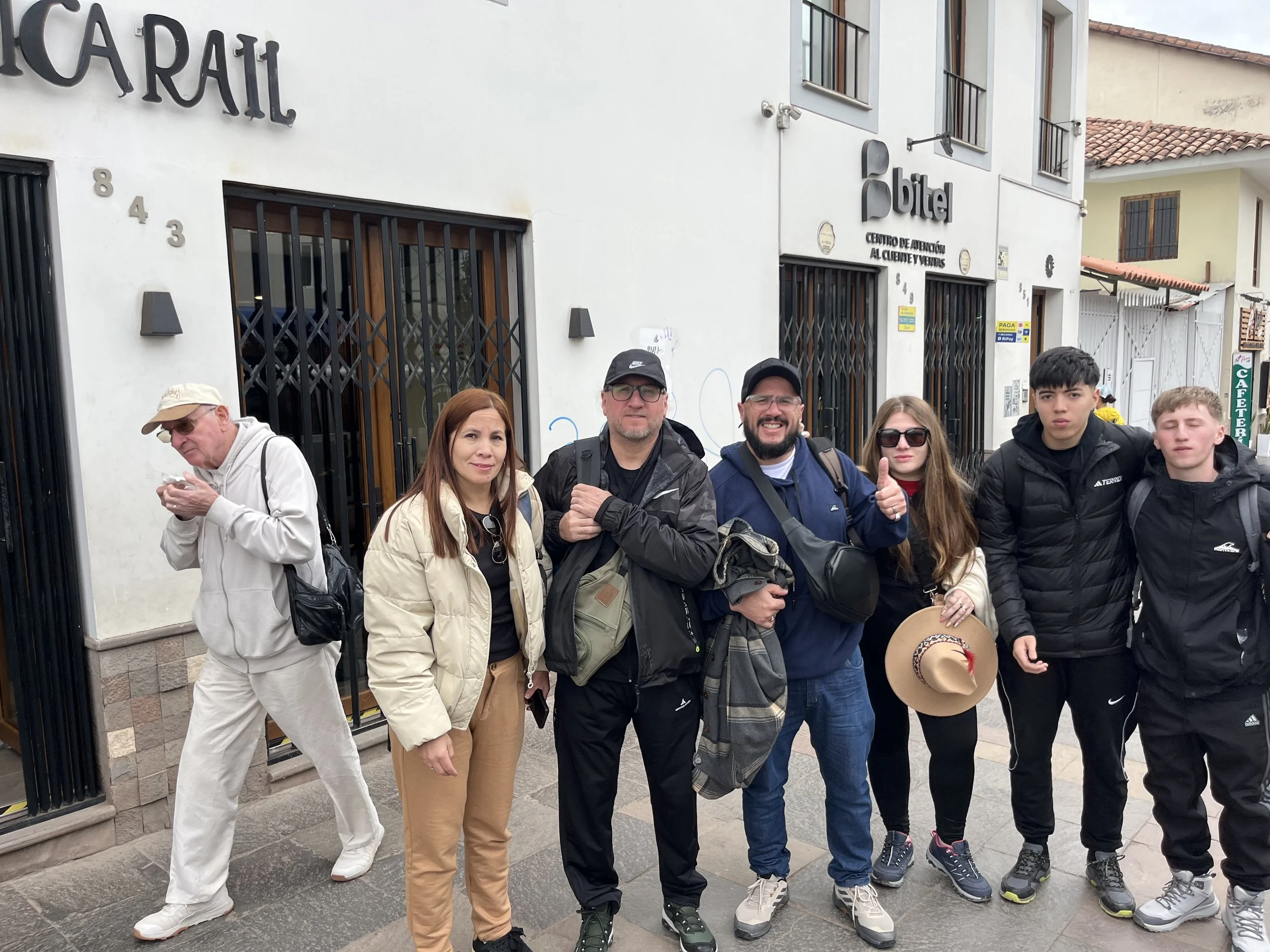 Grupo de personas posando frente a un hotel en una calle urbana, con diferentes edades y estilos de ropa casual y deportiva.