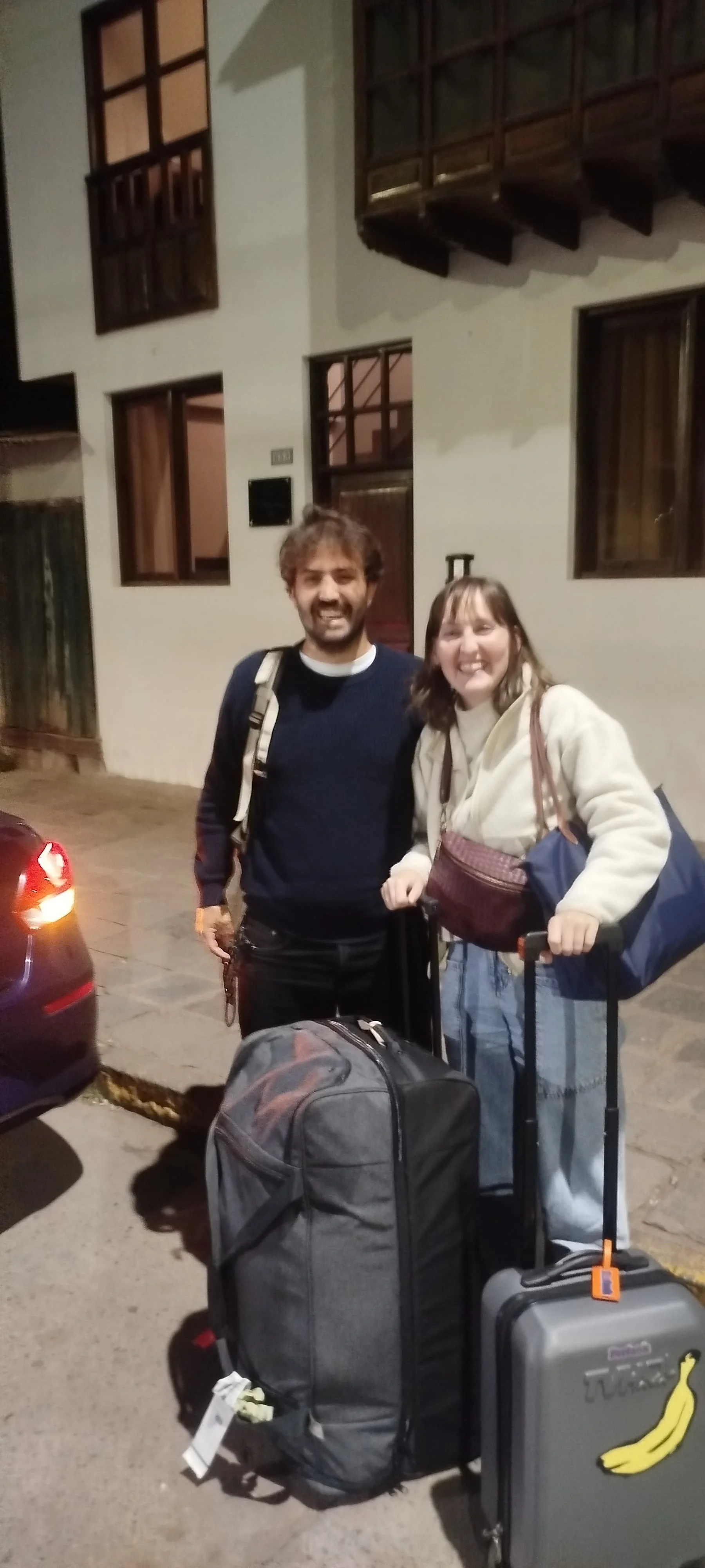 Dos personas de pie en la calle con equipaje, sonriendo. Uno con mochila y la otra con maletín y bolsas, en la noche frente a una casa con ventanas y paredes blancas.