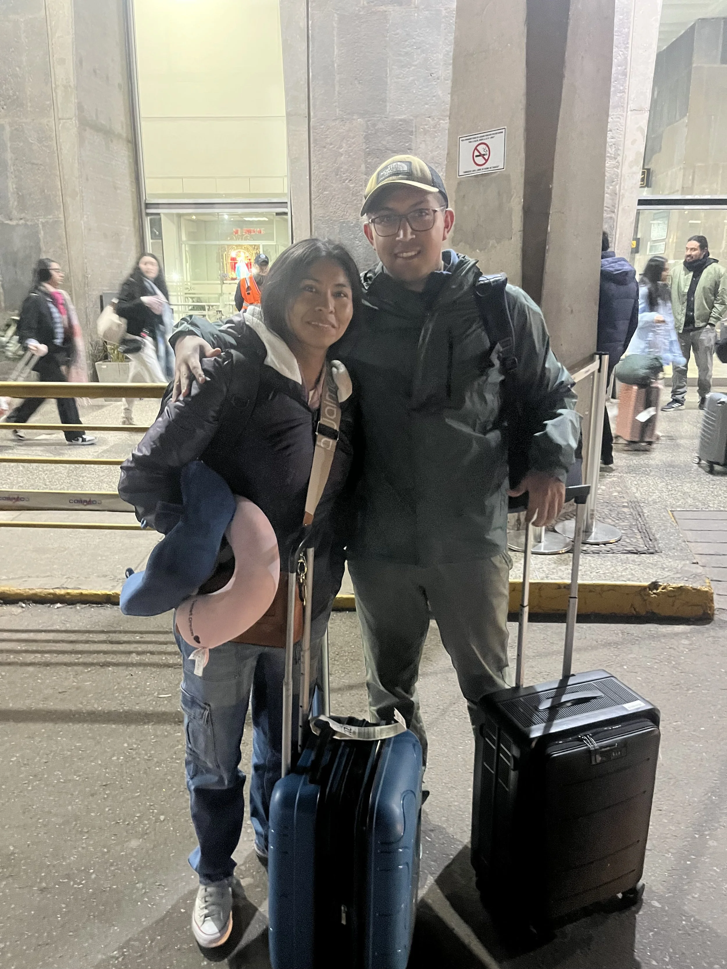 Dos personas con equipaje en la entrada de un aeropuerto por la noche, rodeados de pasajeros.