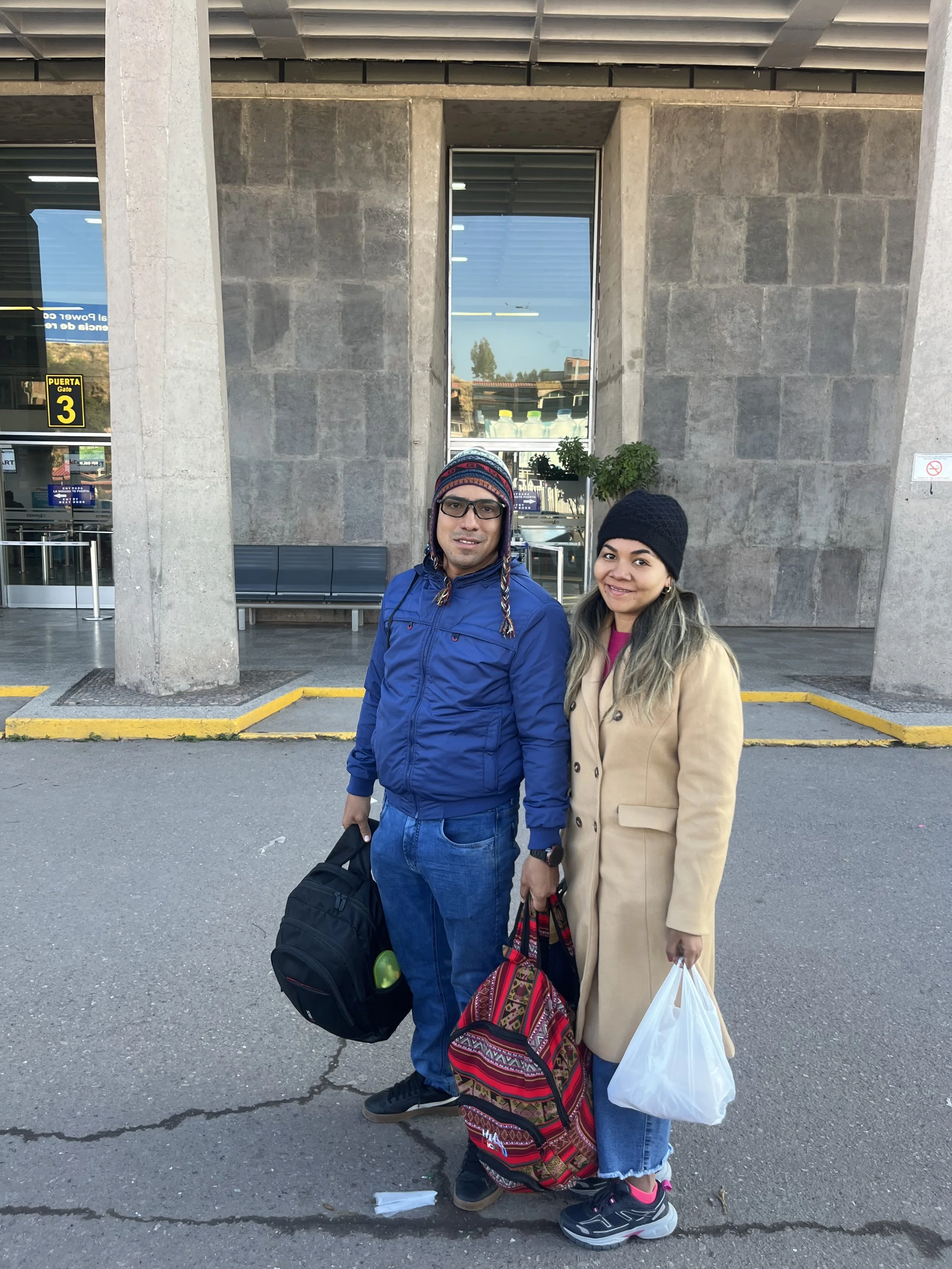 Una pareja con ropa de abrigo y gorros está de pie frente a un edificio, sosteniendo bolsas y mochila, en un entorno urbano.