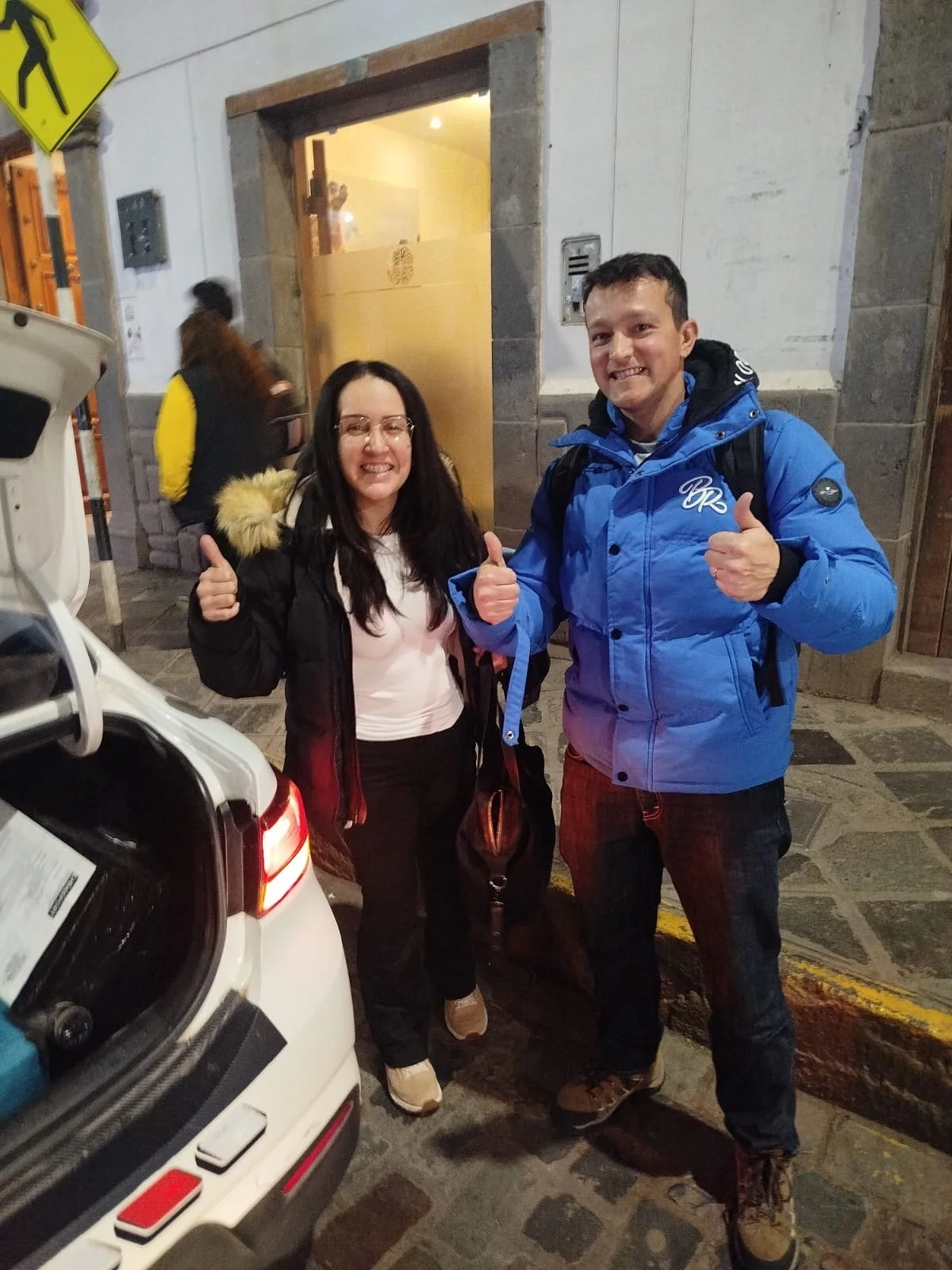 Dos personas, un hombre y una mujer, sonrientes con pulgares arriba, de pie frente a un coche con maletero abierto, en una calle urbana por la noche. Ambos llevan chaquetas abrigadoras.