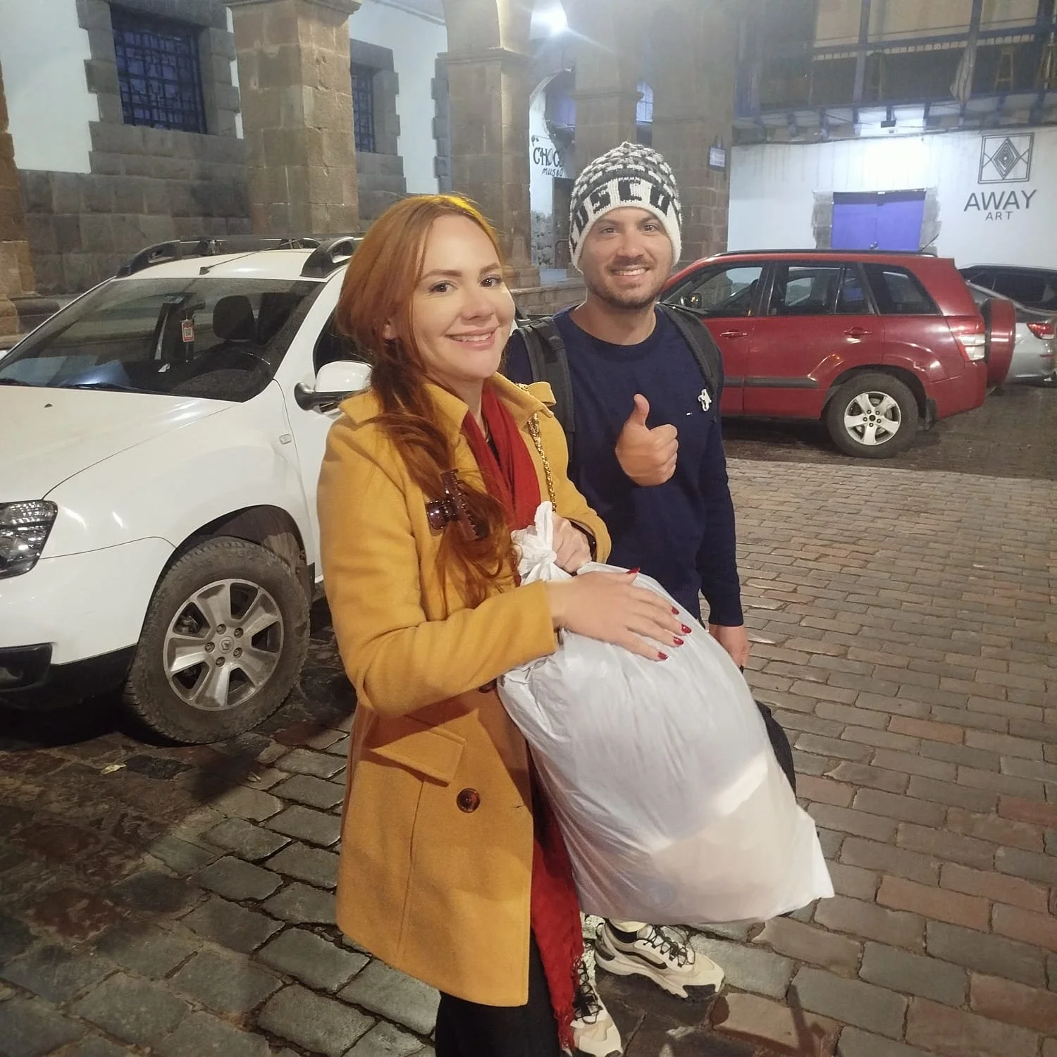 Dos personas, una mujer con abrigo amarillo y una bolsa blanca, y un hombre con gorro y suéter, posando en la calle con autos al fondo.