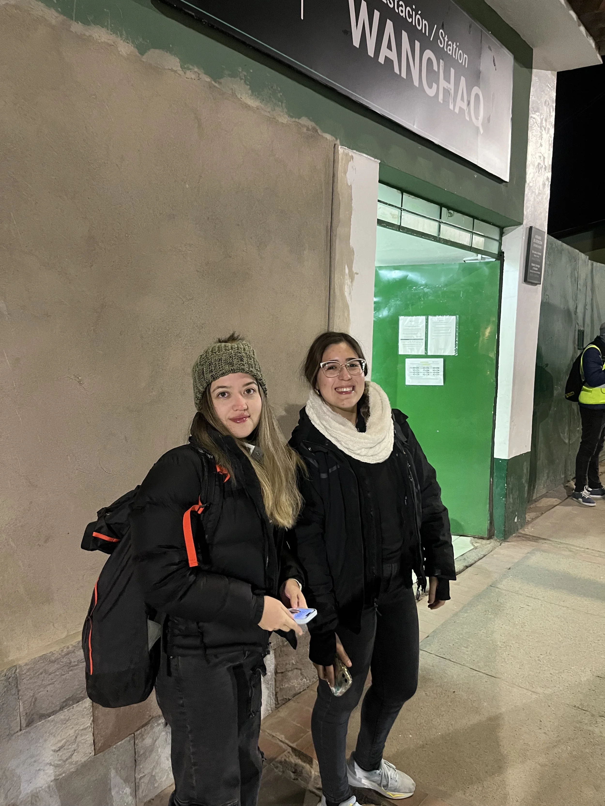 Dos mujeres jóvenes con ropa de abrigo, una con gorro y otra con bufanda, paradas frente a la entrada de la estación Wanchai por la noche.