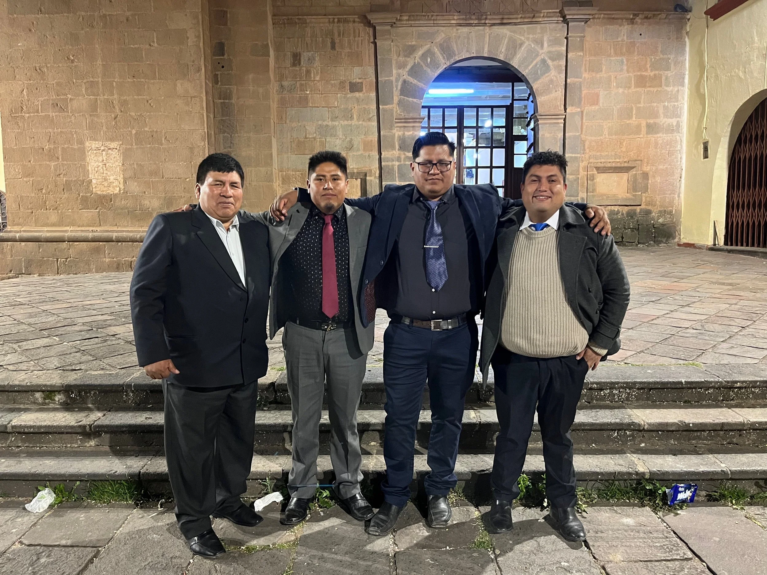 Cuatro hombres jóvenes en vestimenta formal y de negocio, con los brazos entrelazados, posando frente a un edificio de piedra y una ventana con rejas, en la noche.
