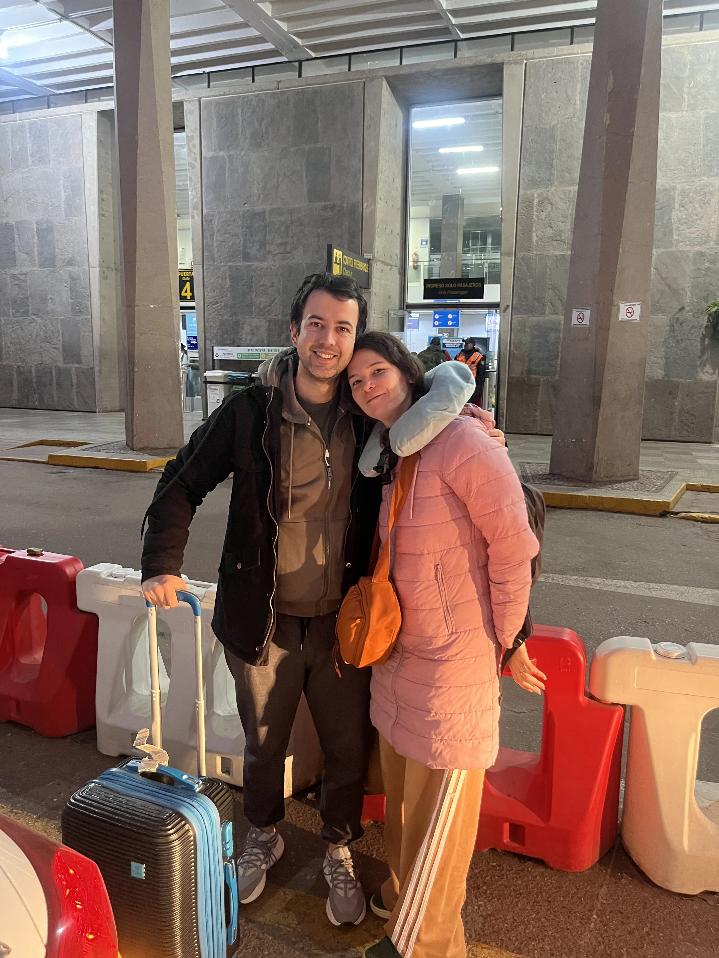 Dos personas felices en un aeropuerto, una con maleta azul y otra con mochila naranja, rodeadas por conos de seguridad
