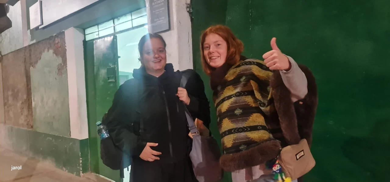 Dos jóvenes sonrientes posando en la calle, una con mochila y otra con bolso y bufanda de colores, en un fondo de pared verde y pared de cemento.
