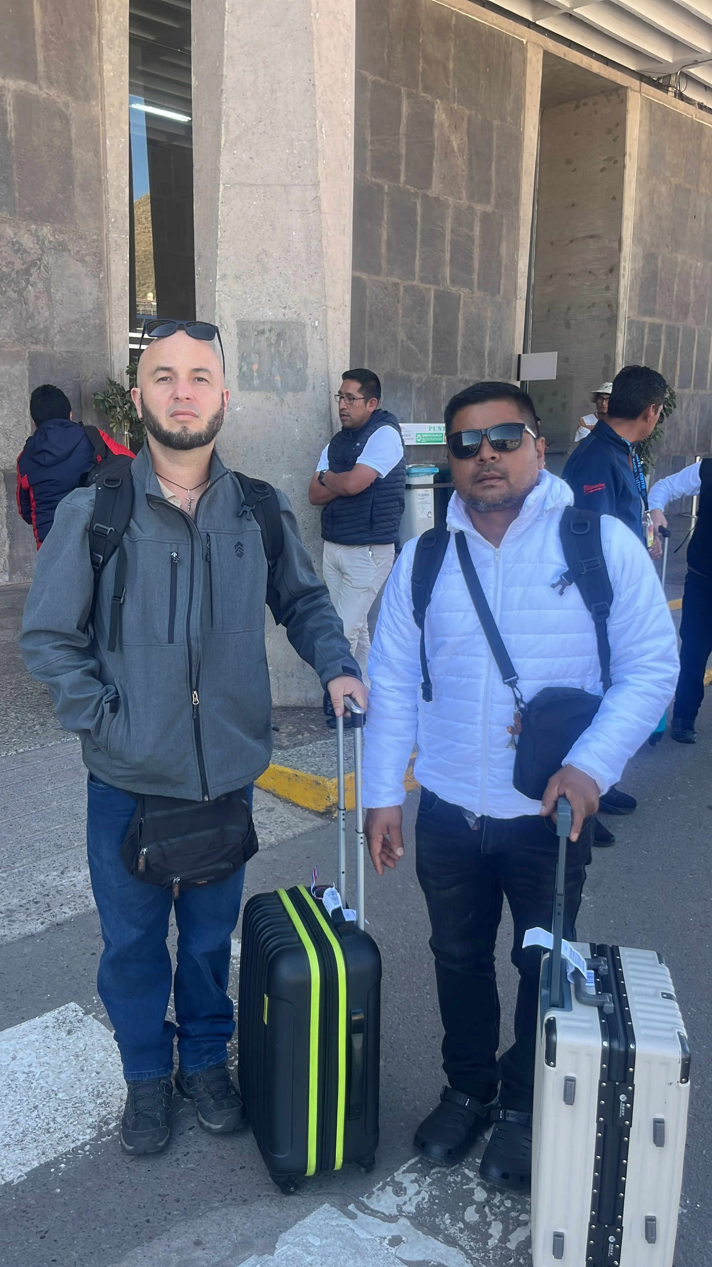 Dos hombres de pie con maletas en una estación de transporte, uno con chaqueta gris y otro con chaqueta blanca, ambos con gafas de sol. Detrás hay otras personas y una estructura de concreto.